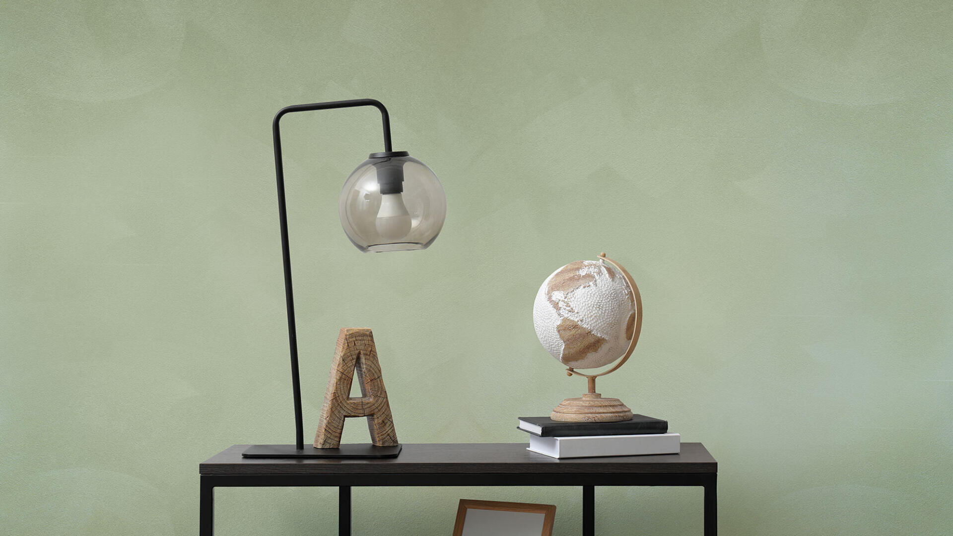 A modern desk with a glass lamp, a decorative letter A wrapped in rope, a neutral-toned globe, two stacked books, and a small picture frame against a light green wall.
