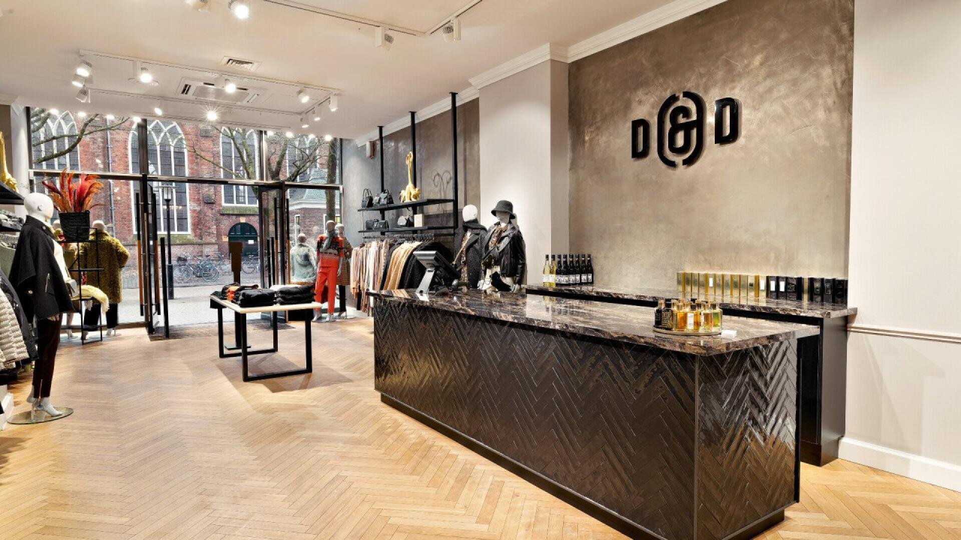 Modern clothing store interior with mannequins dressed in trendy outfits, a marble front counter, clothing racks, and large front windows letting in natural light. The D&D logo is displayed on the textured wall behind the counter.