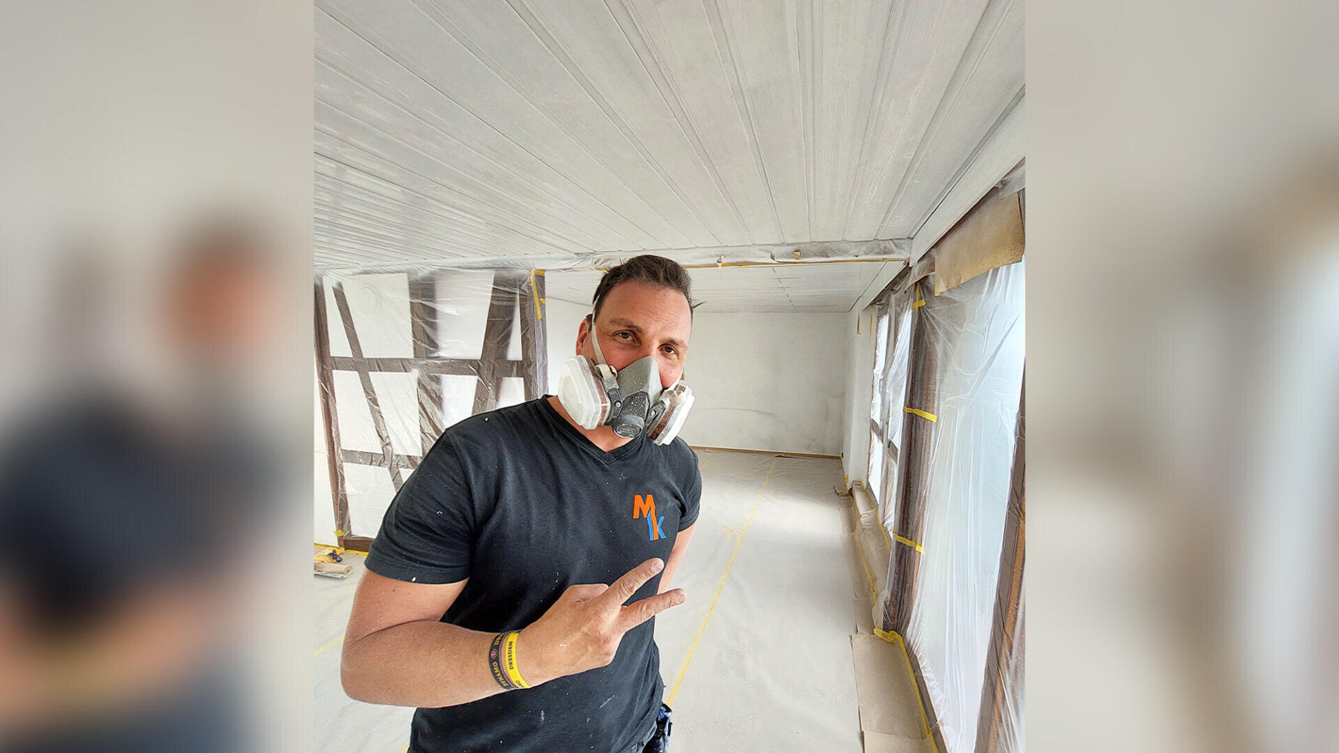 Ein Mann in schwarzem T-Shirt und Atemschutzmaske steht in einem Raum, der gerade renoviert wird, und macht das Peace-Zeichen. Der Raum ist mit schützenden Plastikfolien und Klebeband abgedeckt, die Wände und die Decke sind weiß.