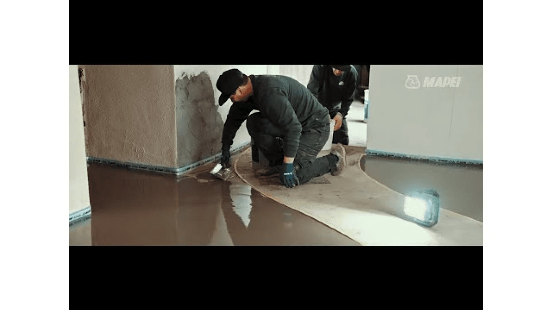 Zwei Arbeiter in dunkler Kleidung glätten nasses Bodenmaterial mit Werkzeugen in der Nähe einer Wand. Sie knien auf Schutzmaterial, während eine Arbeitsleuchte den Bereich beleuchtet. Im Hintergrund ist das Wort MAPEI an der Wand zu sehen.