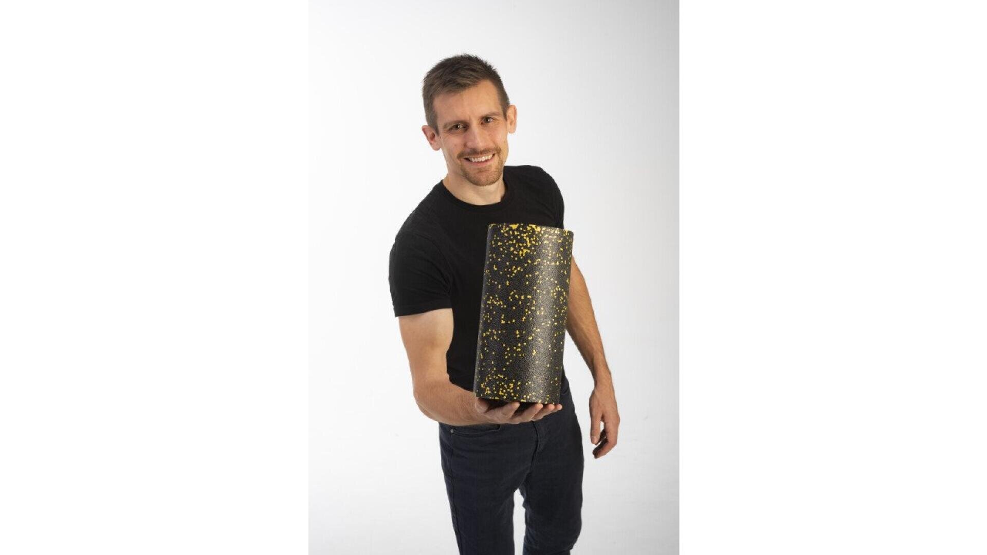 A smiling man in a black t-shirt and jeans holds a black foam roller with yellow specks toward the camera against a plain white background.