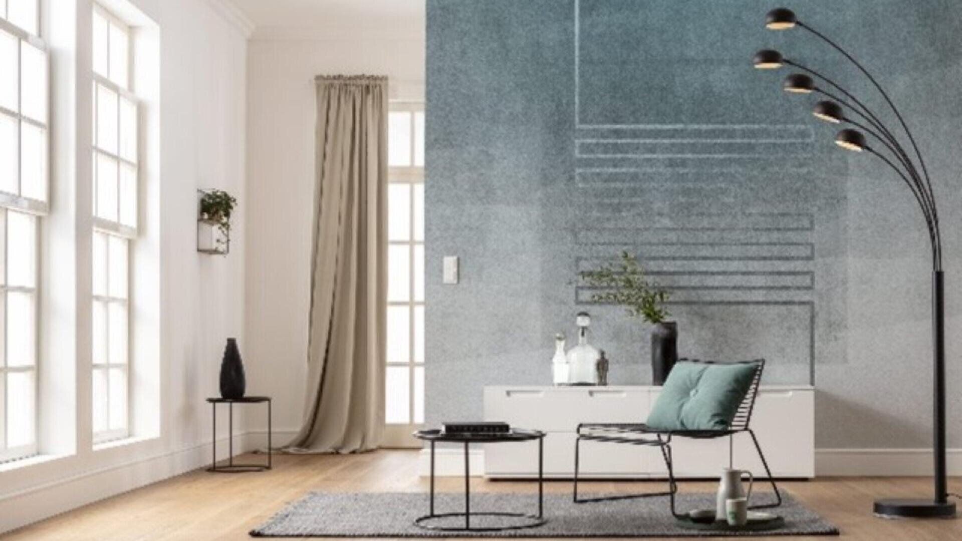 A modern living room with large windows, a gray accent wall, a black floor lamp, a minimalist chair with a green cushion, a small side table, a gray rug, and a white console with decor items.