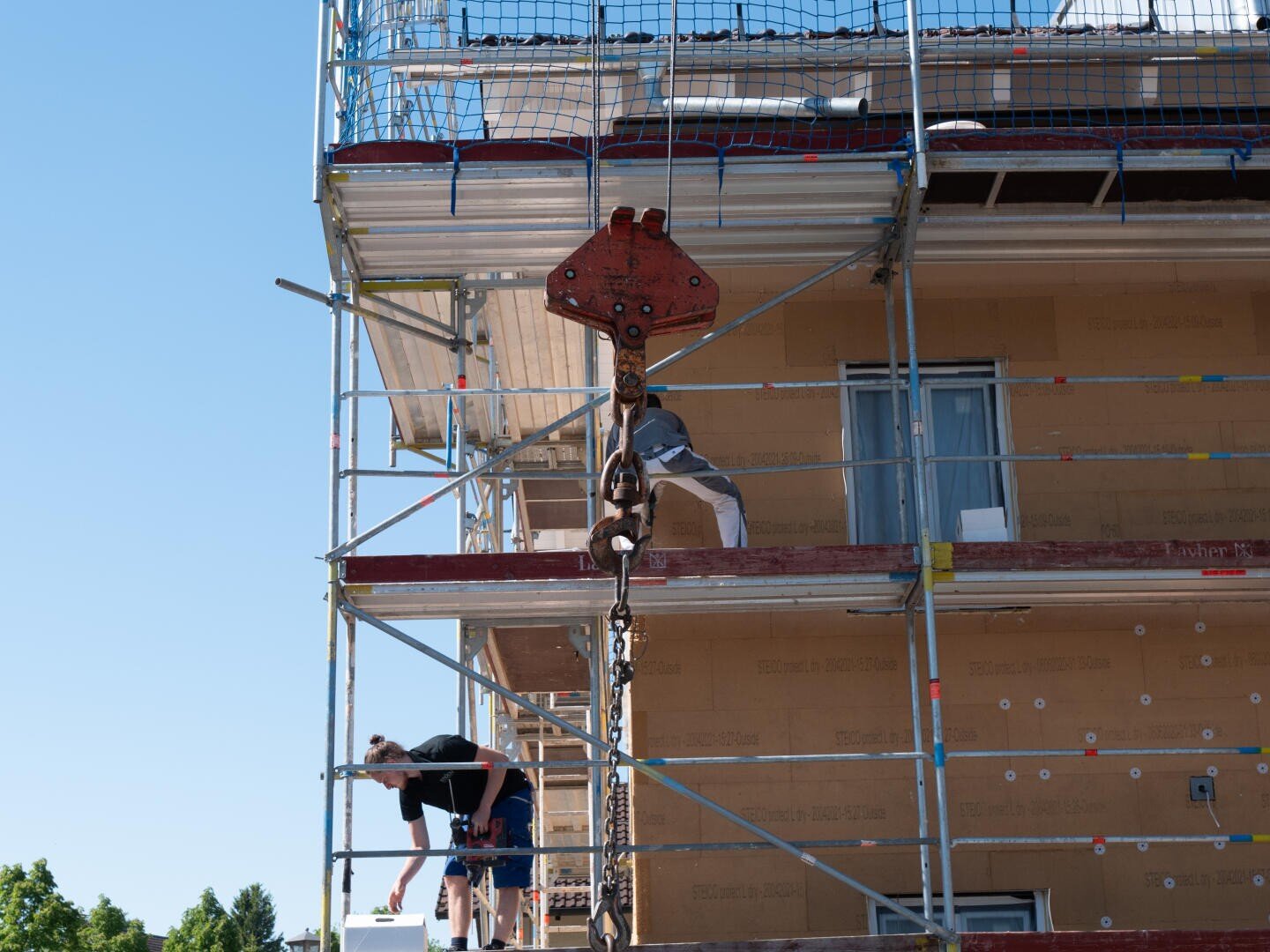 Ein Bauarbeiter steht auf einem Gerüst neben einem im Bau befindlichen Gebäude und hält einen großen Kranhaken und eine Kette fest. Ein anderer Arbeiter arbeitet gebückt auf der unteren Ebene. Im Hintergrund blauer Himmel.