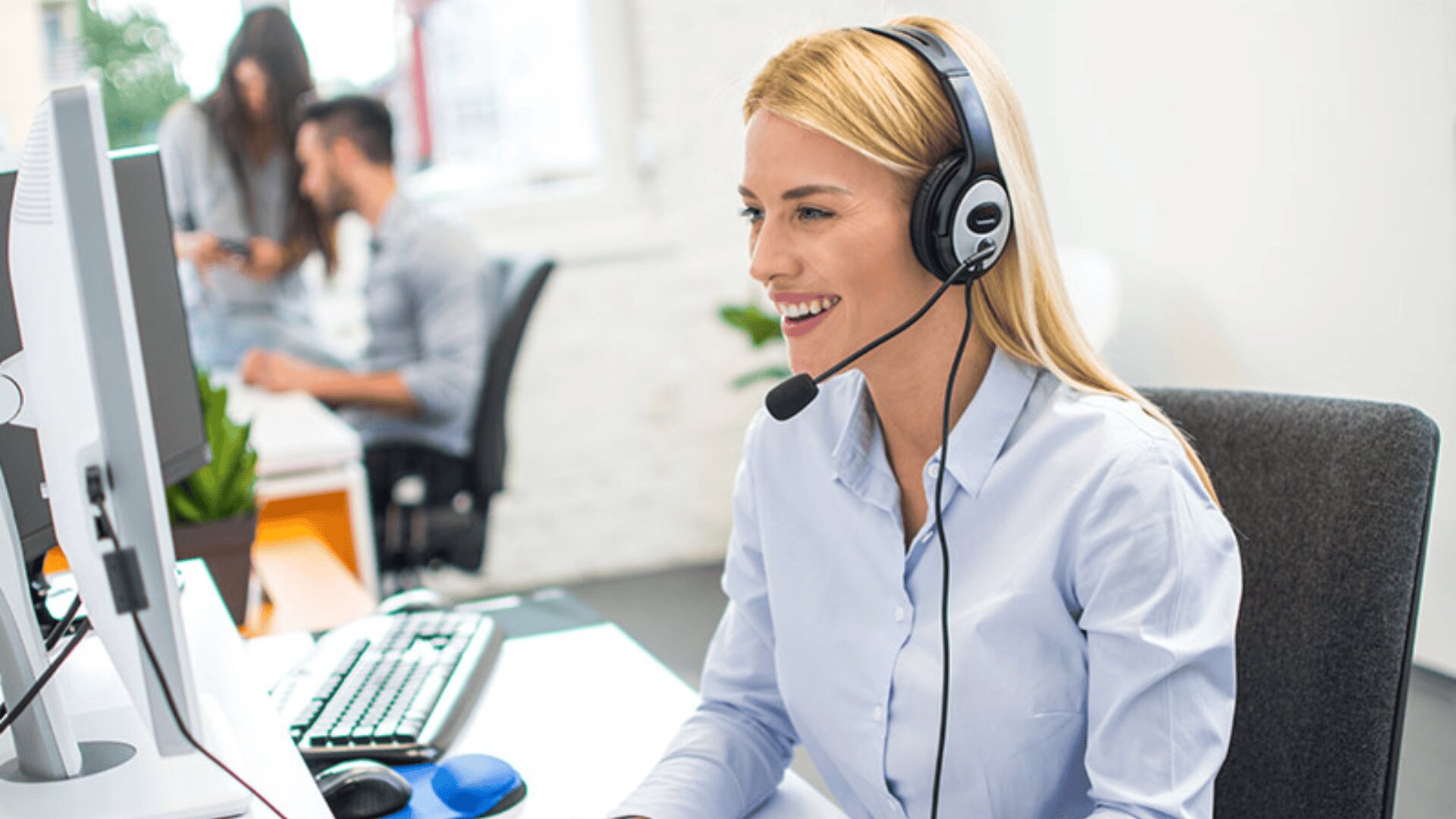 Eine lächelnde Frau mit Headset sitzt an einem Schreibtisch in einem Büro und arbeitet am Computer. Im Hintergrund unterhalten sich zwei Kollegen.