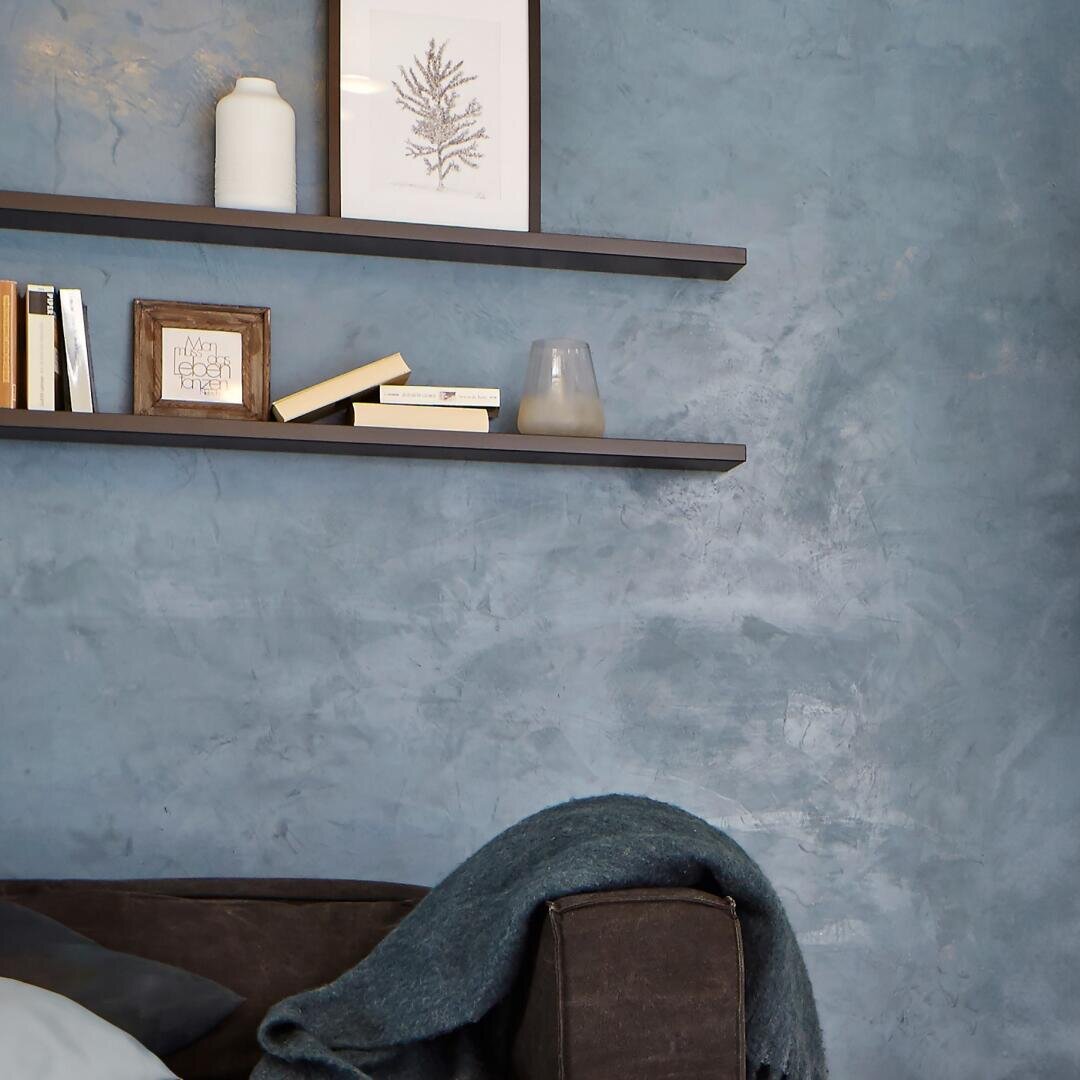 Eine blau strukturierte Wand mit zwei schwebenden Regalen, auf denen Bücher, ein gerahmtes Bild, eine weiße Vase und ein Glas stehen. Im Vordergrund ist die Ecke eines braunen Sofas zu sehen, über dessen Armlehne eine dunkle Decke hängt.