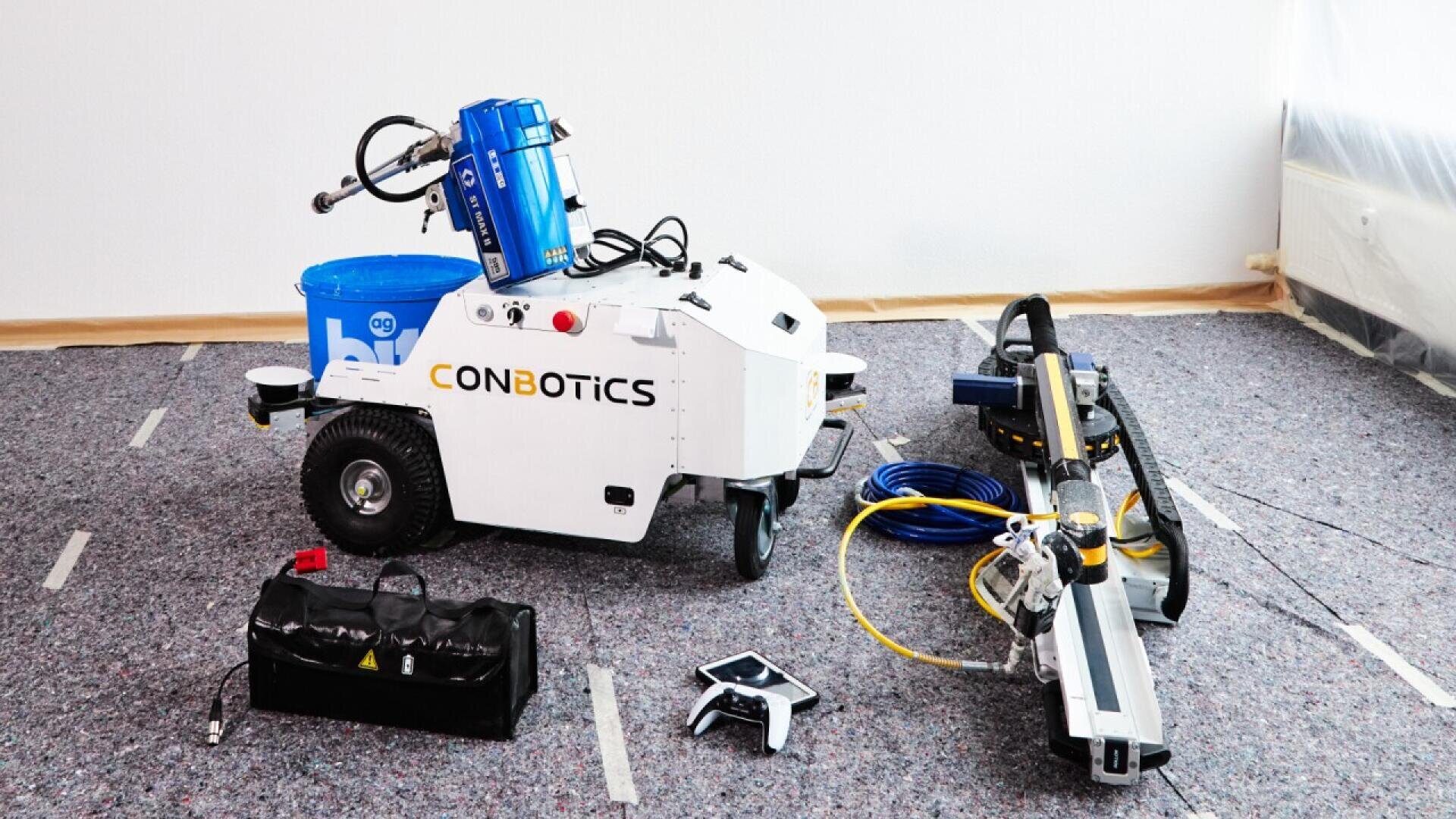 A white robot labeled CONBOTICS with attached blue equipment and cables is positioned on a carpeted floor, surrounded by technical gear, a black case, and other machinery components. The area appears to be a construction or tech setup.