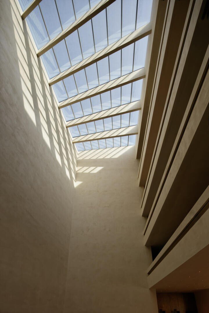 Hohe, moderne Innenansicht eines Gebäudes mit einem schrägen Glasdach, das Sonnenlicht hereinlässt und Schatten auf beige Wände wirft, und mehreren parallelen Balkonen auf der rechten Seite.