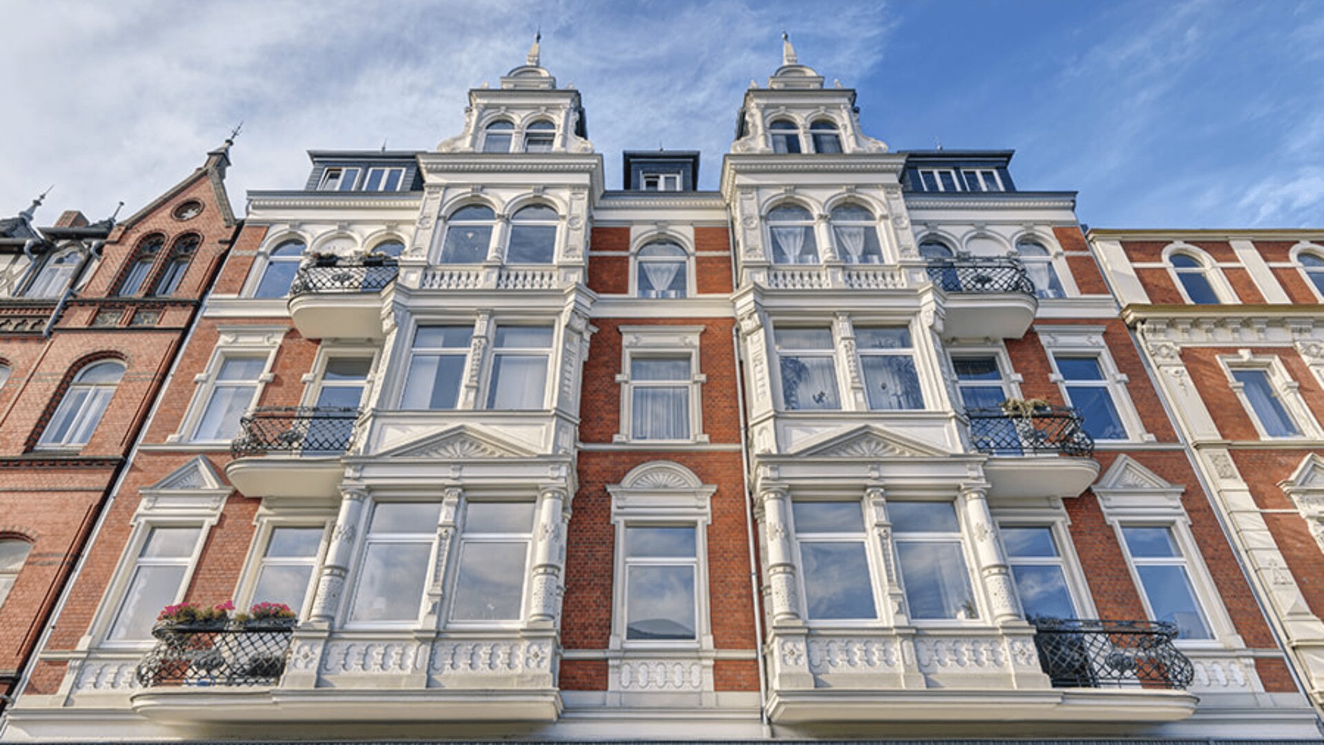 Elegantes historisches Gebäude mit verzierter Fassade aus weißen und roten Ziegeln, hohen Fenstern, Balkonen mit schwarzem Geländer und dekorativen architektonischen Details unter einem blauen Himmel mit vereinzelten Wolken.