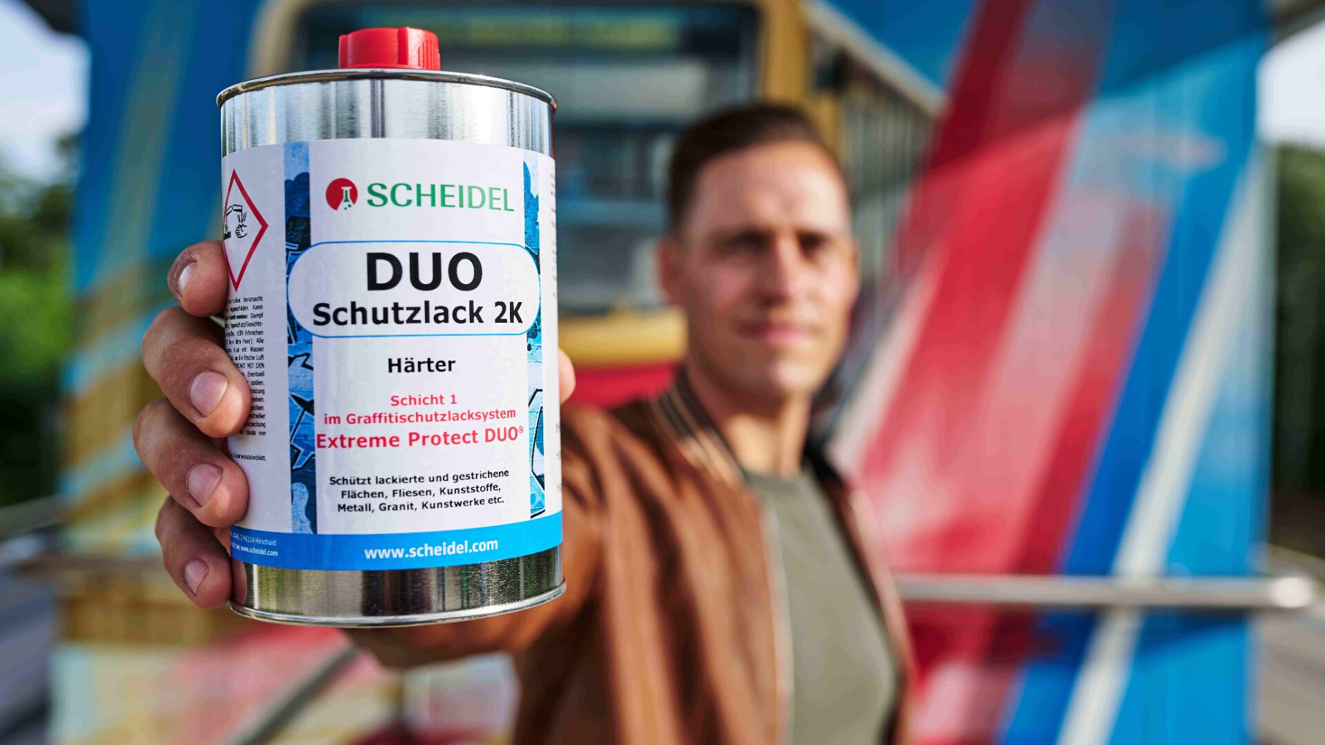 Eine Person hält eine Dose Scheidel DUO Schutzlack 2K in die Kamera, dahinter sind bunte abstrakte Formen und ein unscharfer Hintergrund zu sehen.