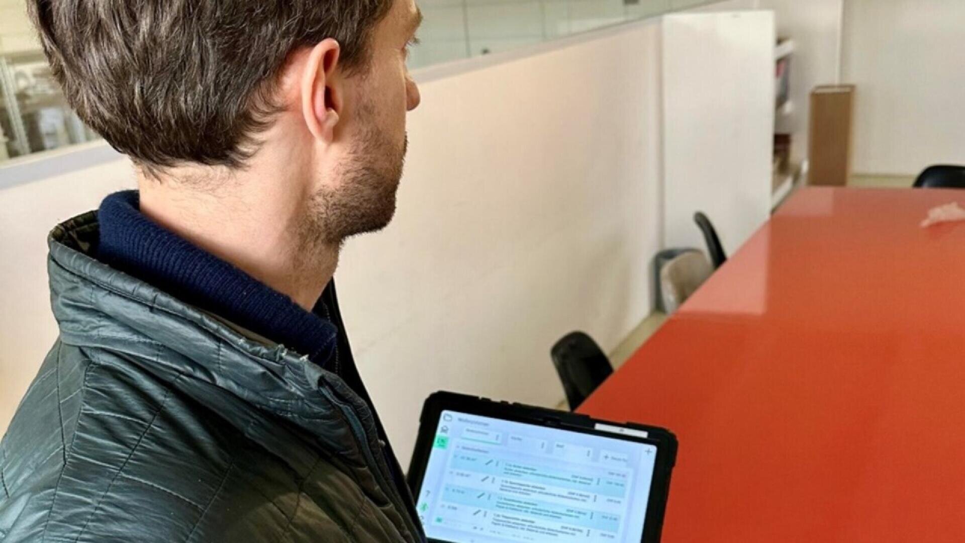 A person holding a tablet with a visible screen stands next to a long red conference table in a modern office space.