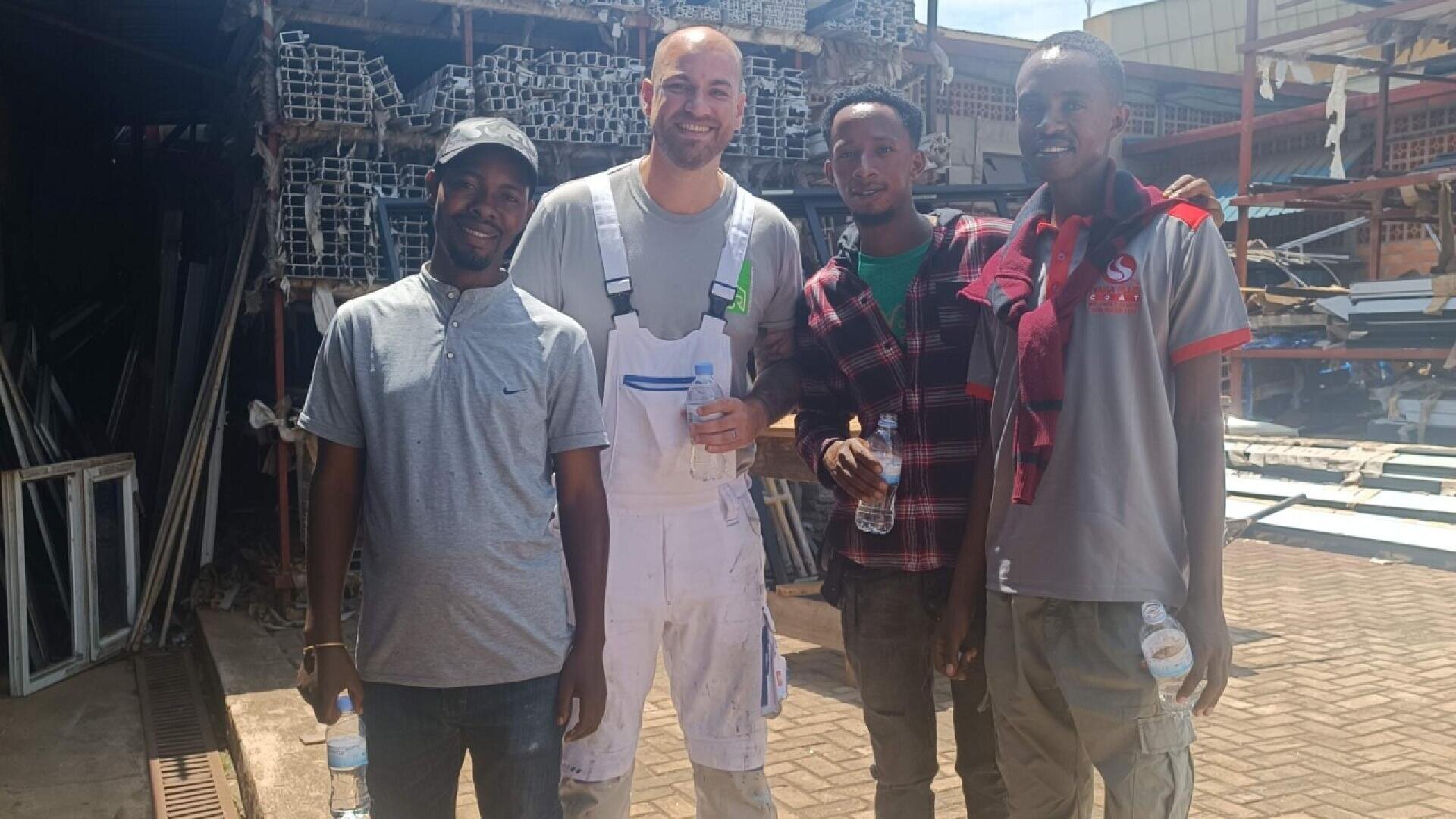 Four men stand together smiling at a construction site with metal materials stacked in the background. The man second from left wears white overalls, while the others wear casual shirts and pants.
