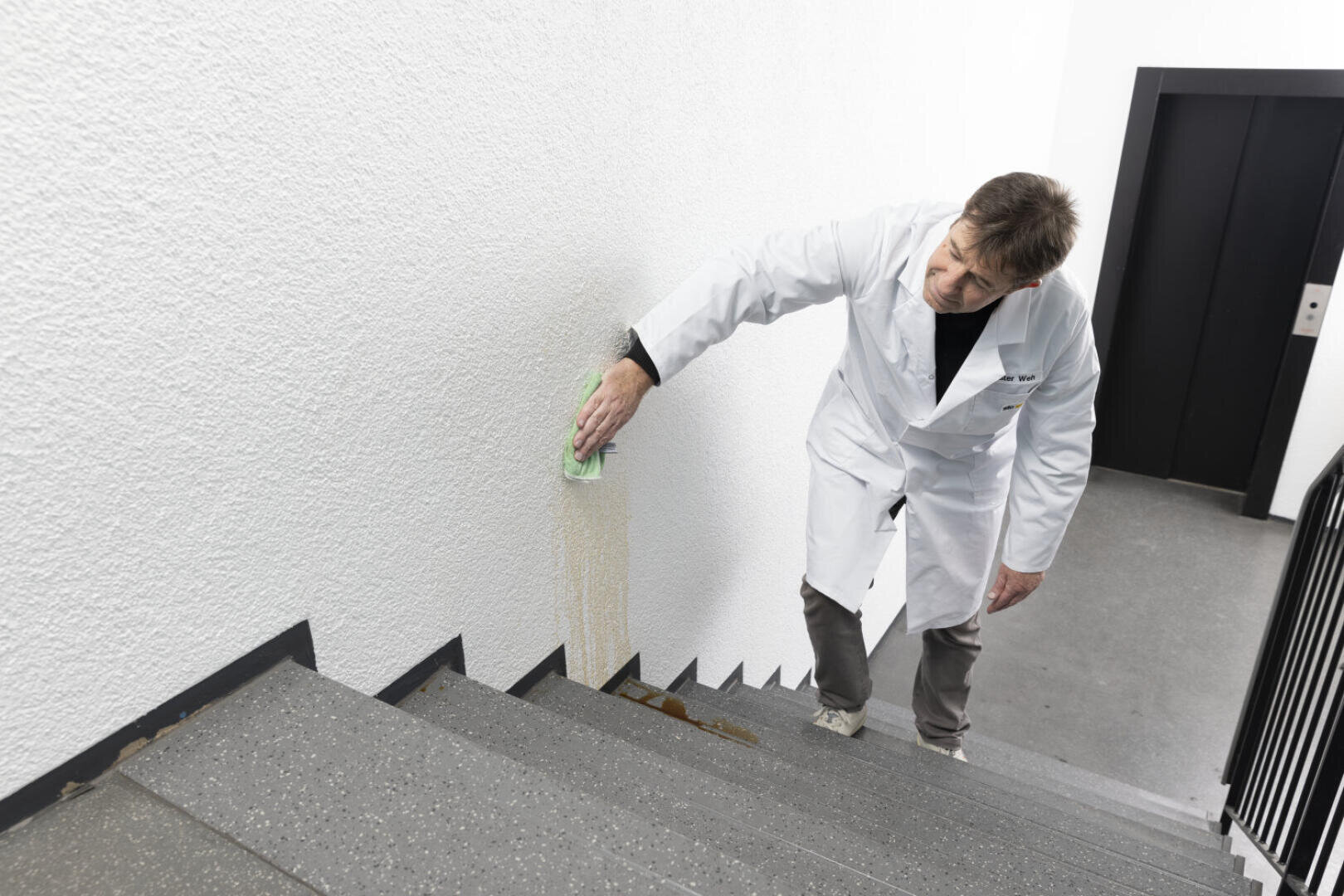 Ein Mann im weißen Laborkittel wischt mit einem grünen Tuch eine verschüttete Flüssigkeit an der Wand neben einer Treppe ab. Er steht auf der Treppe neben einem schwarzen Handlauf und einer geschlossenen Aufzugstür.