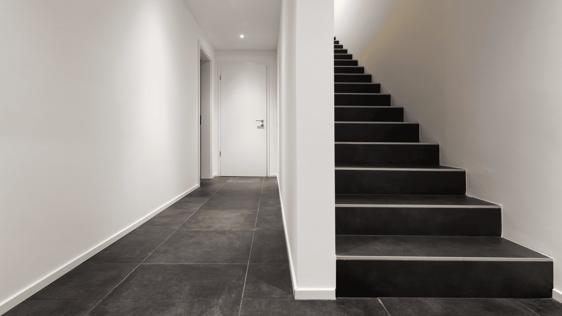 Ein minimalistischer Flur mit dunklem Fliesenboden, weißen Wänden, einer weißen Tür am Ende und einer Treppe mit schwarzen Stufen und weißen Setzstufen auf der rechten Seite. Der Raum ist sauber, modern und gut beleuchtet.