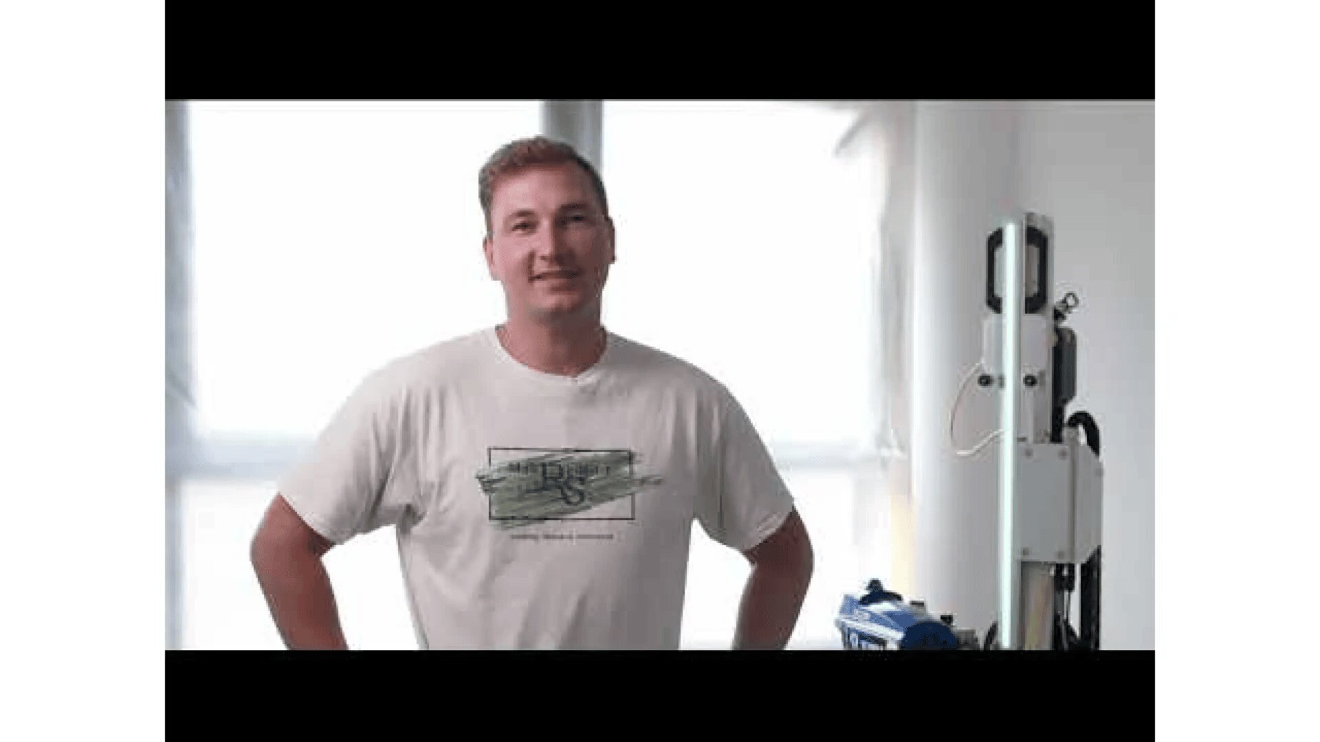 Ein Mann in einem weißen T-Shirt steht mit den Händen in den Hüften im Innenbereich und lächelt in die Kamera. Auf der rechten Seite des Bildes ist Ausrüstung, möglicherweise für Fotografie oder Technik, zu sehen.
