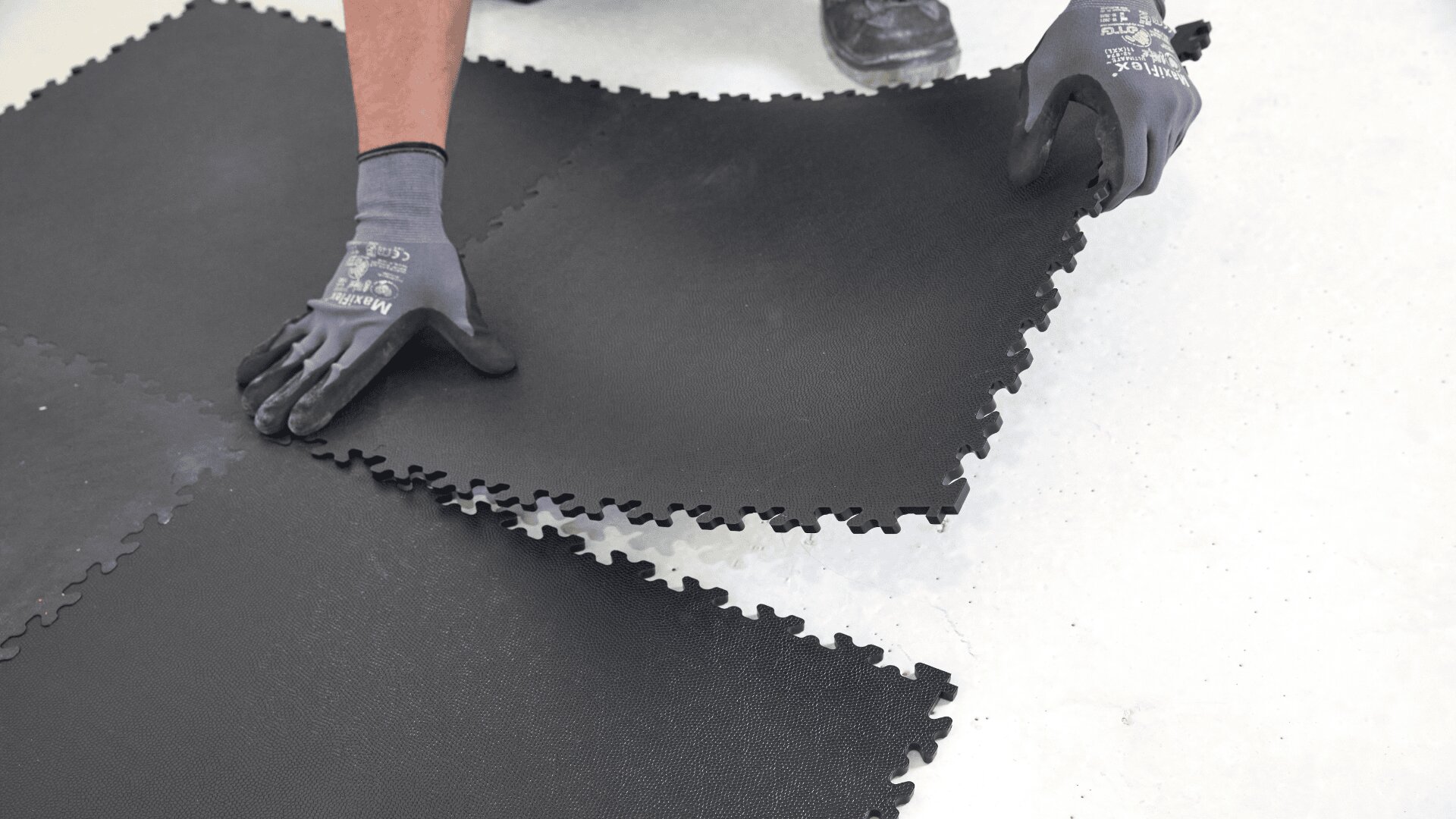 Eine Person mit schwarzen Handschuhen fügt große schwarze, ineinandergreifende Schaumstoff-Bodenfliesen auf einer hellen Oberfläche zusammen.