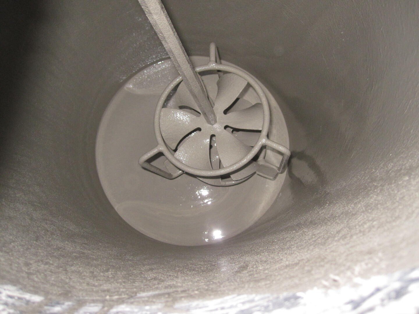 Blick von oben auf einen industriellen Mischtank mit einem Metallrührer im Inneren, umgeben von einer gräulichen Aufschlämmung oder Flüssigkeit, die den Boden und die unteren Wände des Tanks bedeckt.