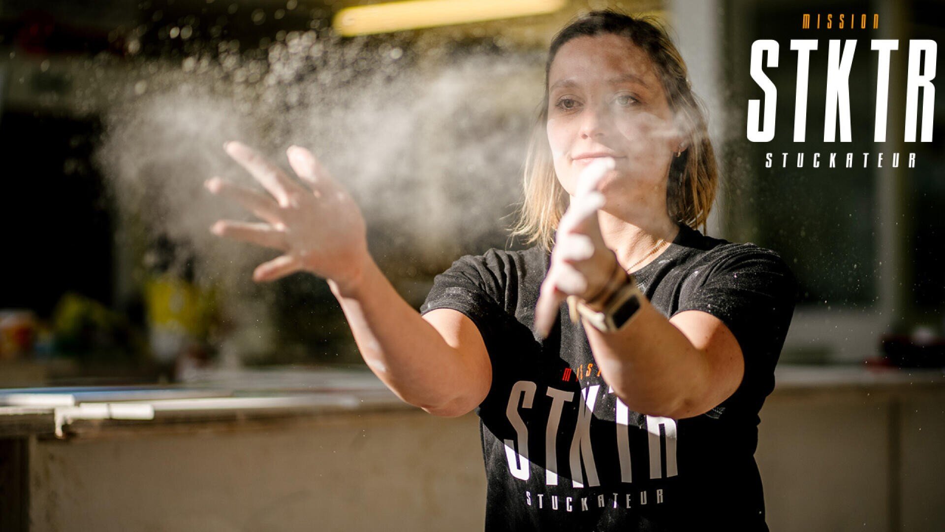 Eine Frau in einem schwarzen STKTR-Shirt wirft weißes Pulver in die Luft, das vom Sonnenlicht beleuchtet wird. Das STKTR-Logo erscheint in der oberen rechten Ecke des Bildes.