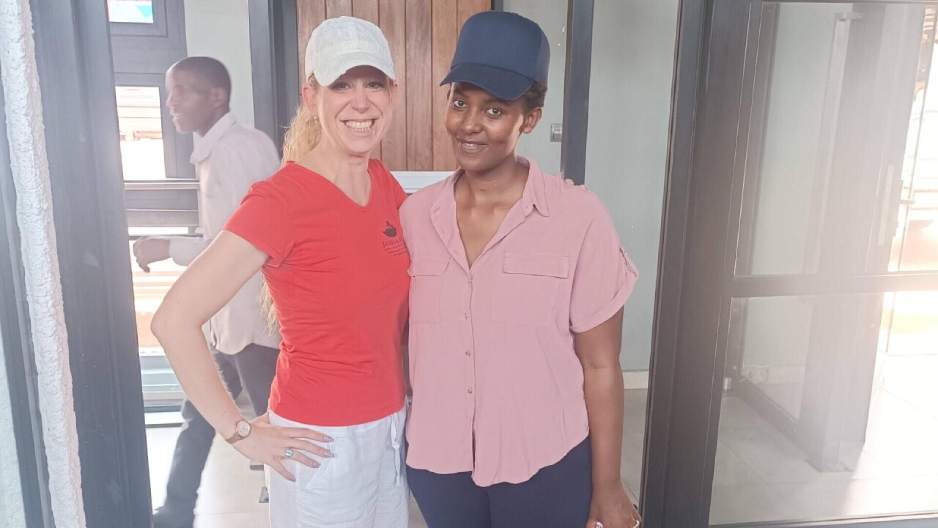 Two women stand side by side indoors, smiling at the camera. One wears a red shirt, white pants, and a light cap; the other wears a pink shirt, dark pants, and a dark cap. A person walks by in the background.