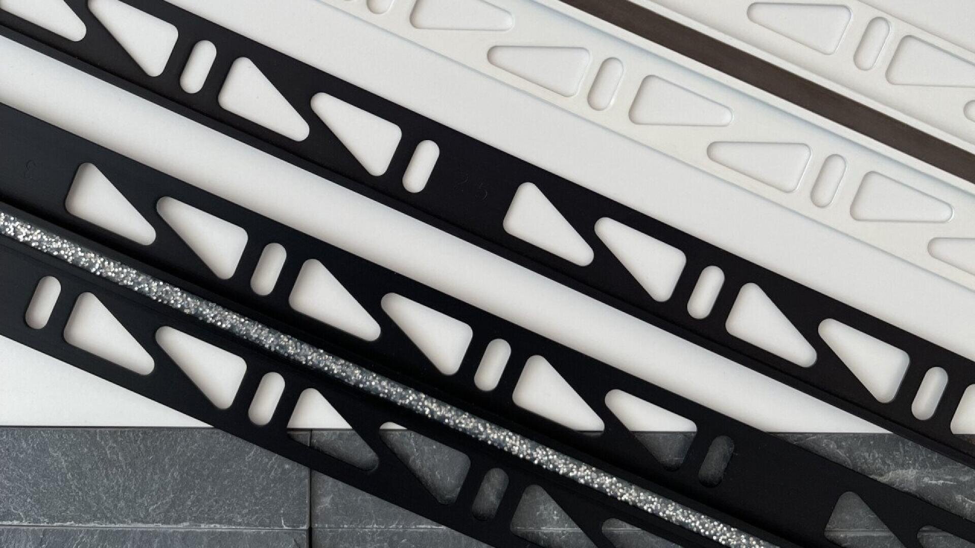 Diagonal view of several metal or plastic strips with geometric triangular and rectangular cutouts, placed on a tile and white background. The strips are black, white, or silver, with one black strip featuring a glittery texture.