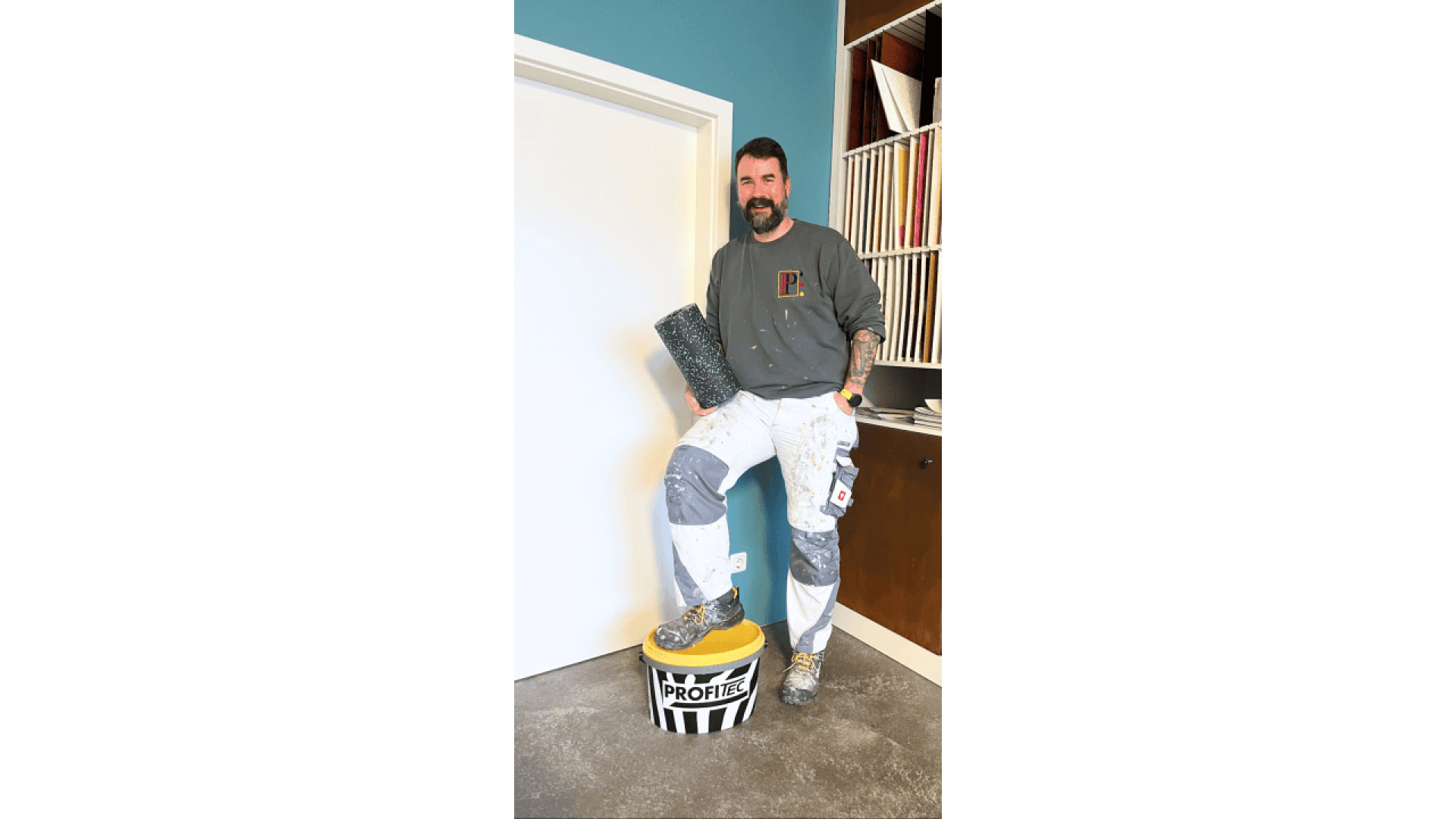 A bearded man in a gray shirt and paint-splattered pants stands with one foot on a yellow paint bucket, holding a textured roller, in a room with a blue wall and shelves with files.