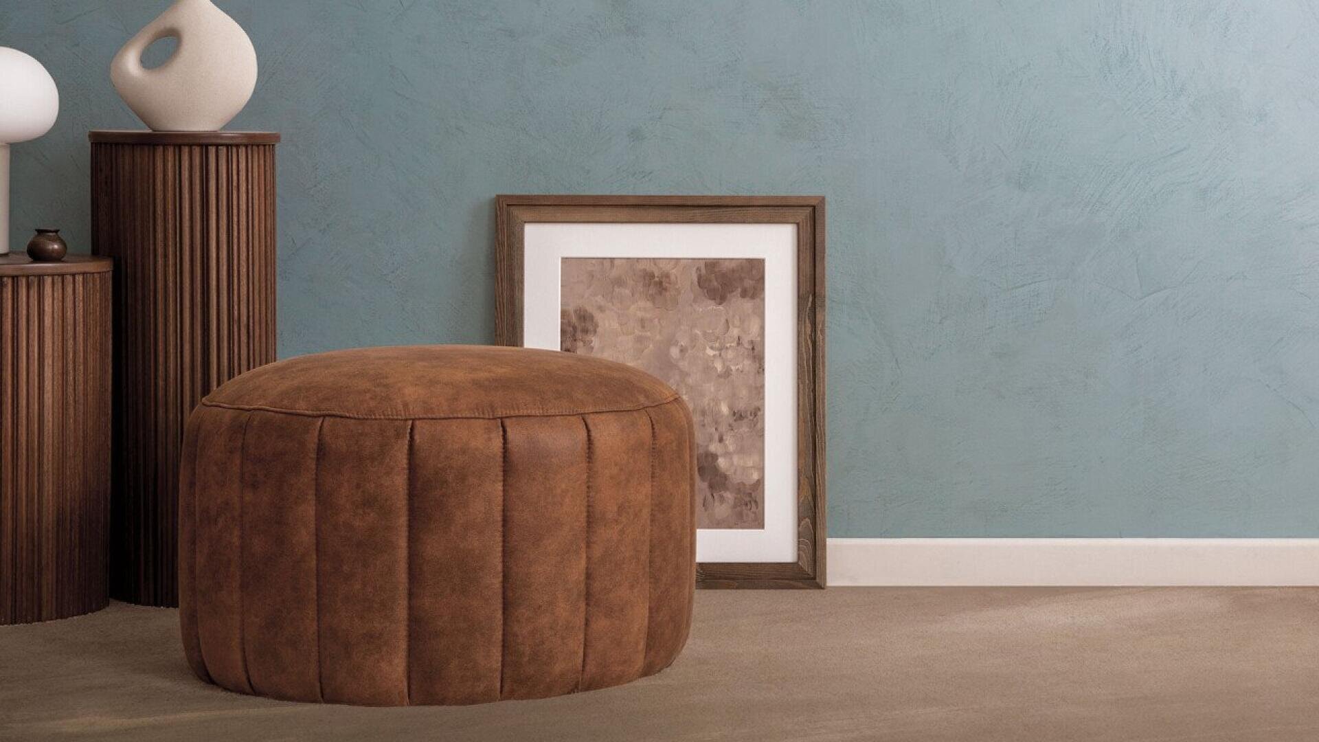 Ein brauner Lederpouf steht auf einem beigen Boden neben zwei gerippten Holzsockeln mit Keramikvasen und einem gerahmten abstrakten Kunstwerk, das an einer blau strukturierten Wand lehnt.