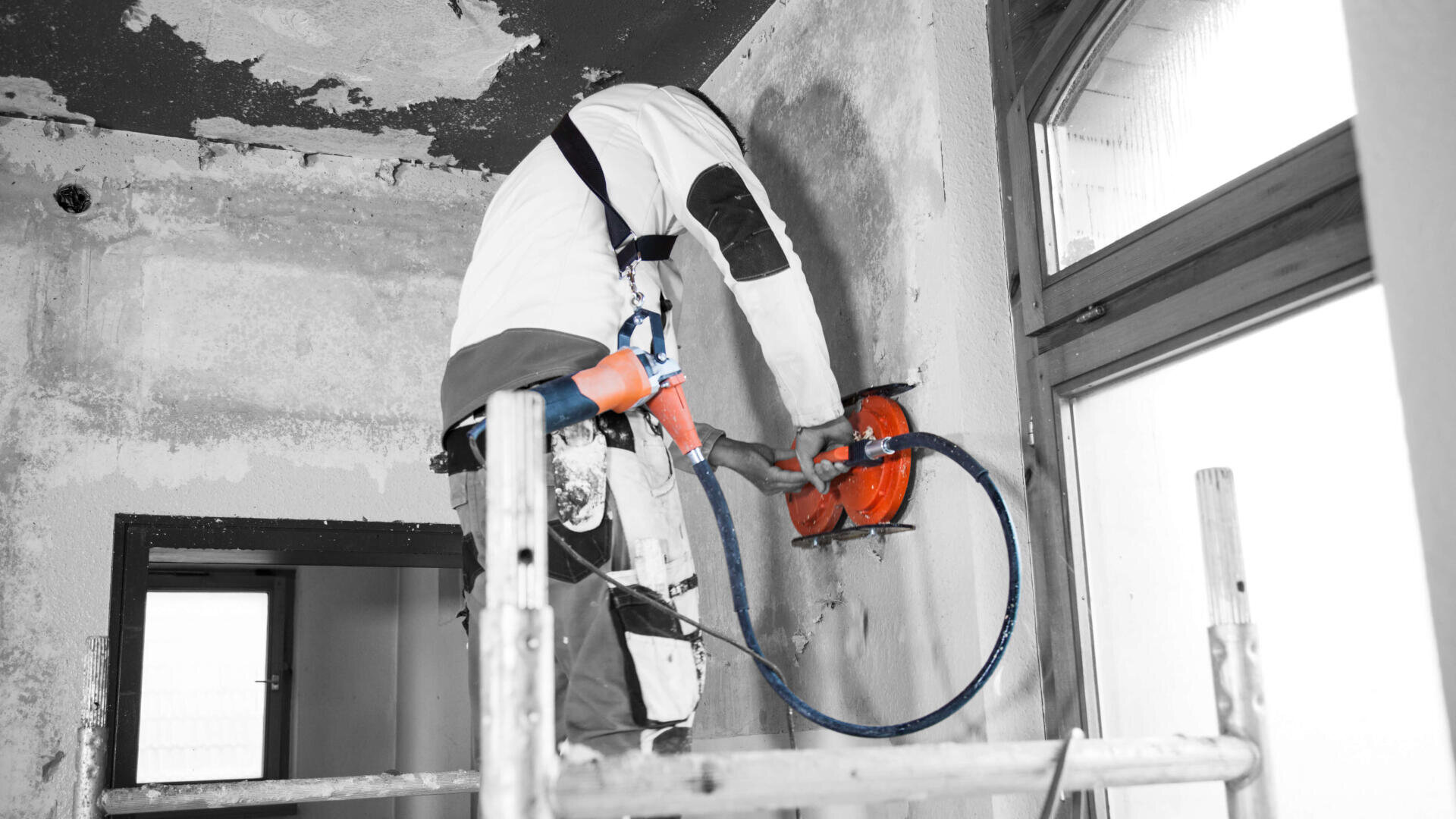 Ein Arbeiter in Schutzausrüstung steht auf einem Gerüst und bohrt oder schneidet mit einem Elektrowerkzeug in eine Wand in einem Gebäude, das gerade renoviert wird.