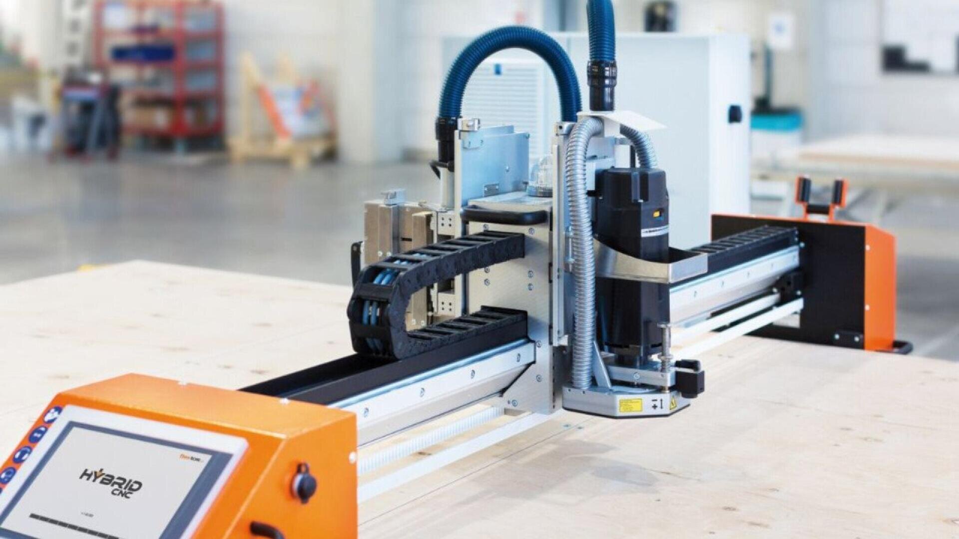 A modern CNC machine with an orange control panel and various mechanical components is set up in a spacious industrial workshop with equipment in the background.