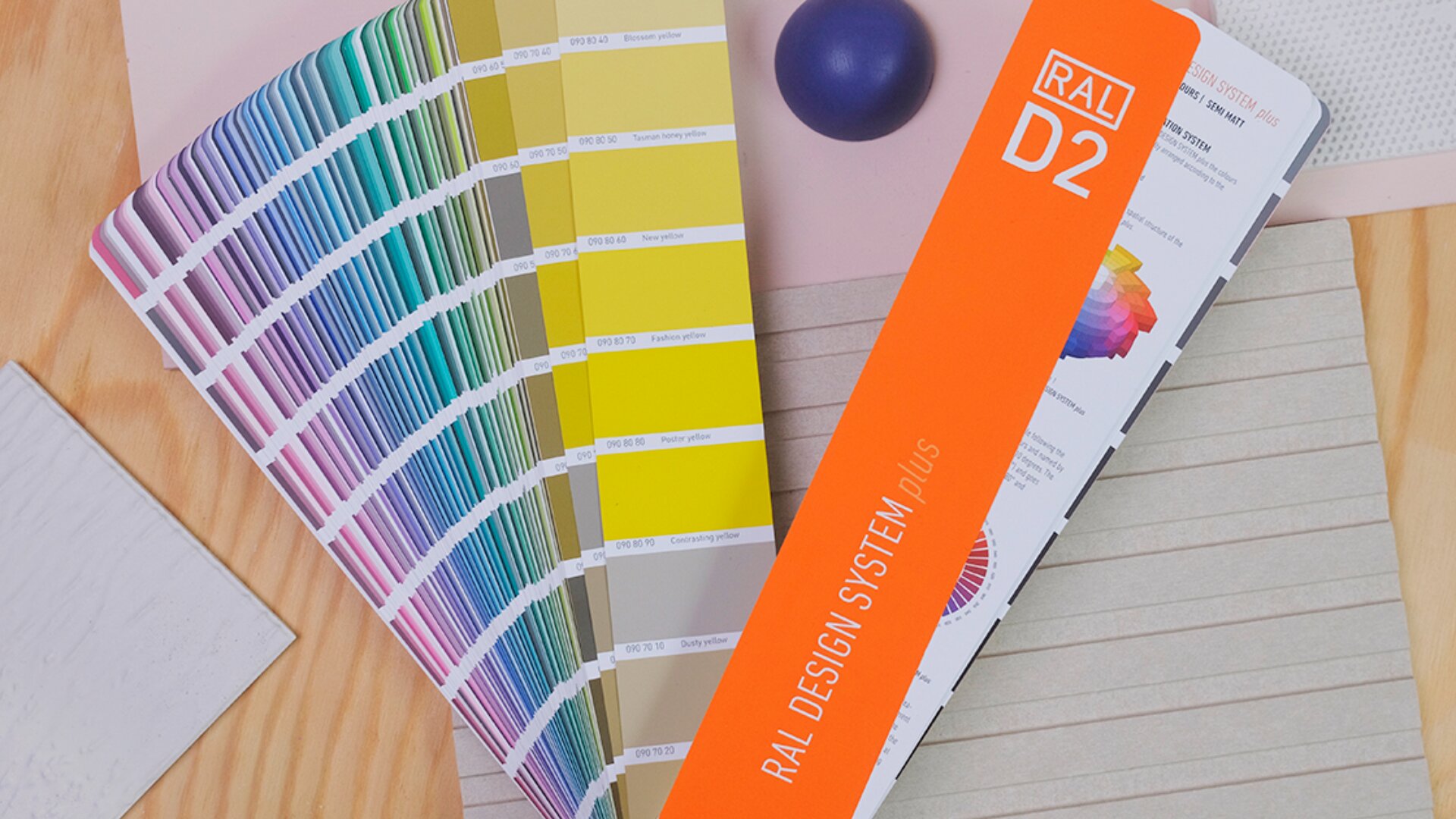 Auf einer Holzoberfläche ist ein Farbfächer des RAL Design Systems Plus mit verschiedenen Farbmustern ausgebreitet. Daneben befindet sich ein orangefarbener Deckel mit der Aufschrift „RAL D2“ und eine violette Kugel.