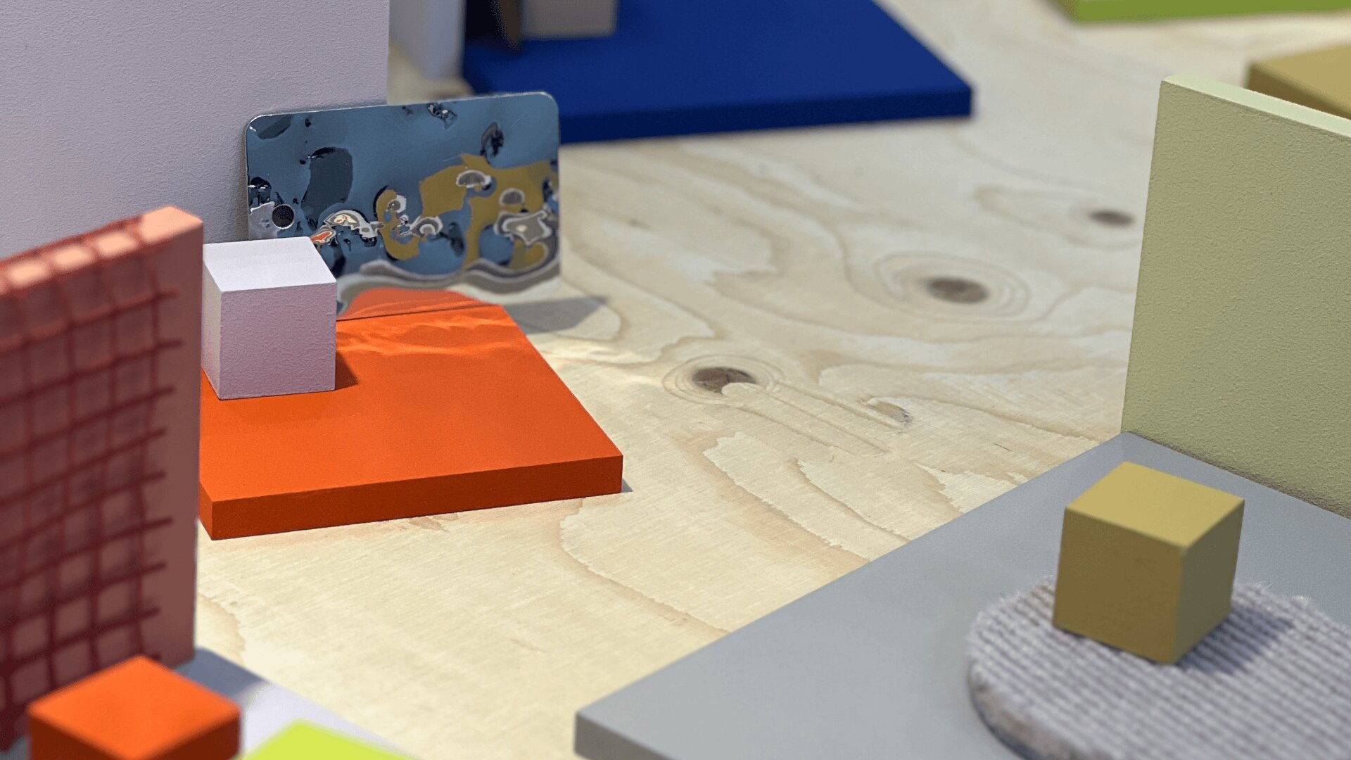 Eine Nahaufnahme einer Holzoberfläche mit bunten Blöcken, Metallteilen und strukturierten Objekten, die in einem abstrakten, geometrischen Muster angeordnet sind. Die Szene enthält orange, blaue, gelbe, graue und weiße Elemente.