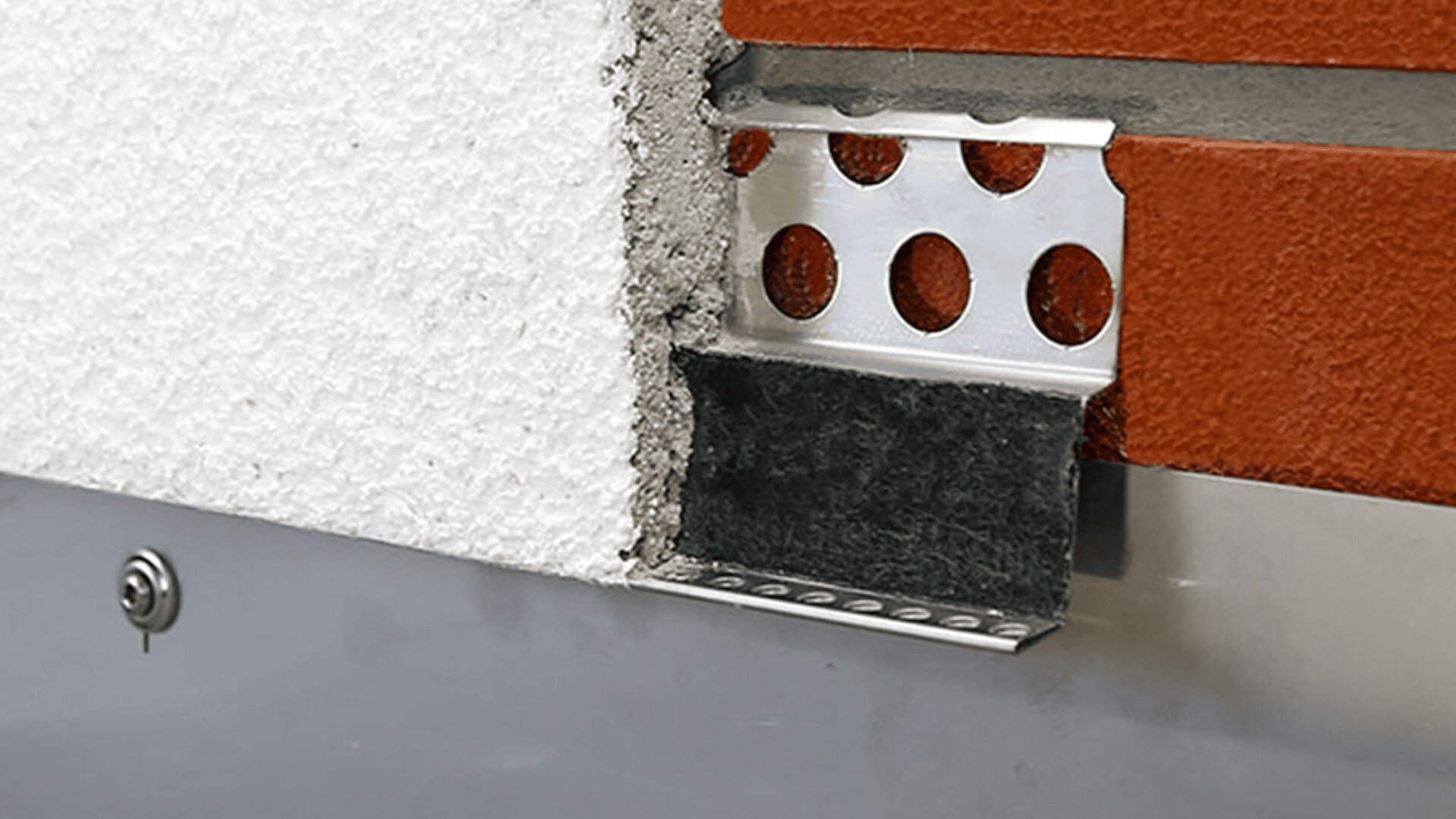 Nahaufnahme eines Gebäudewandquerschnitts mit geschichteten Materialien, darunter weißem Putz, einem Metallprofil mit runden Löchern, einer schwarzen Membran und rotem Mauerwerk, die Konstruktionsdetails veranschaulichen.