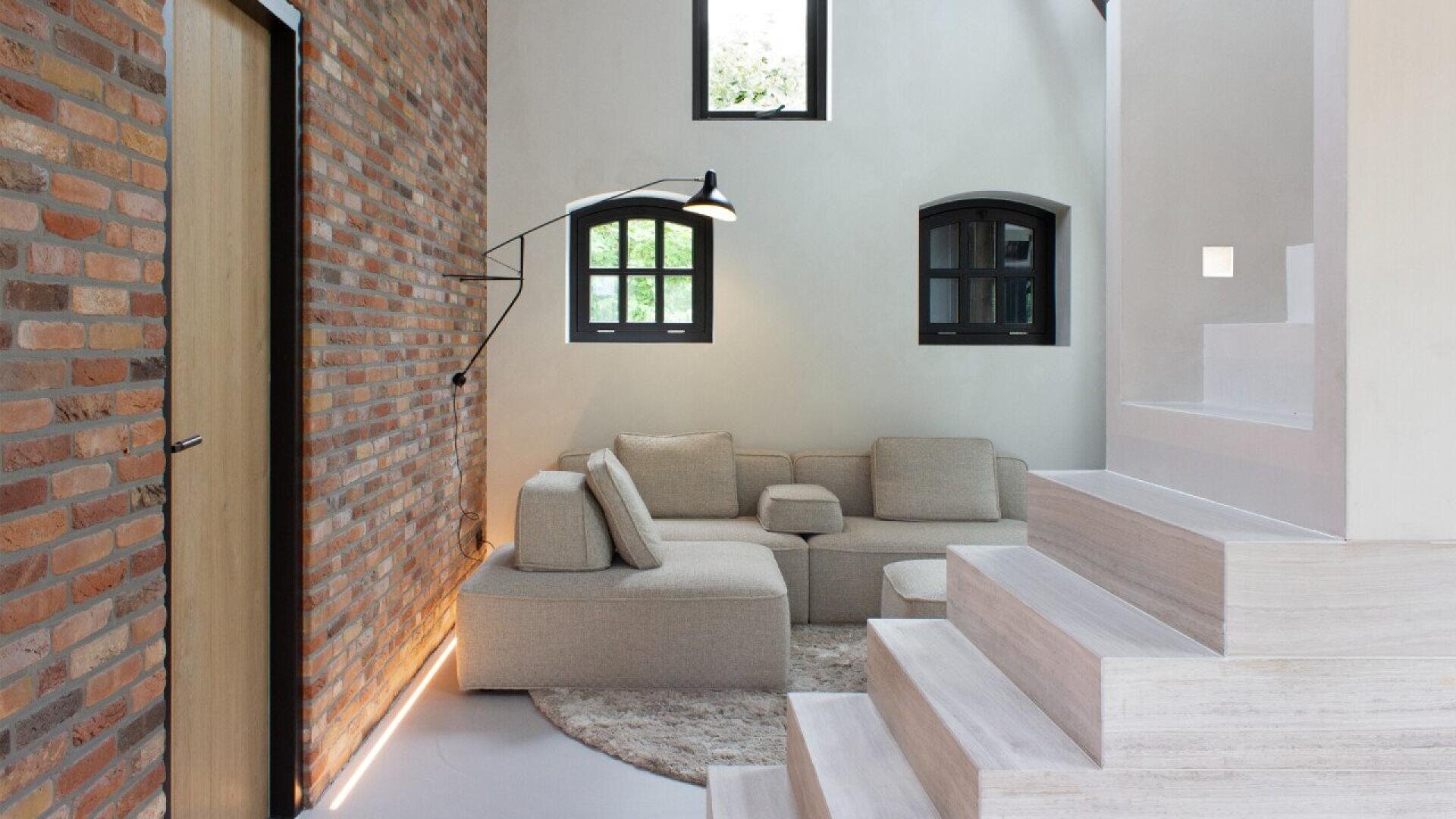 A modern living room with exposed brick walls, a beige sectional sofa on a round rug, black-framed windows, a wall-mounted lamp, and white stairs leading up to another level.