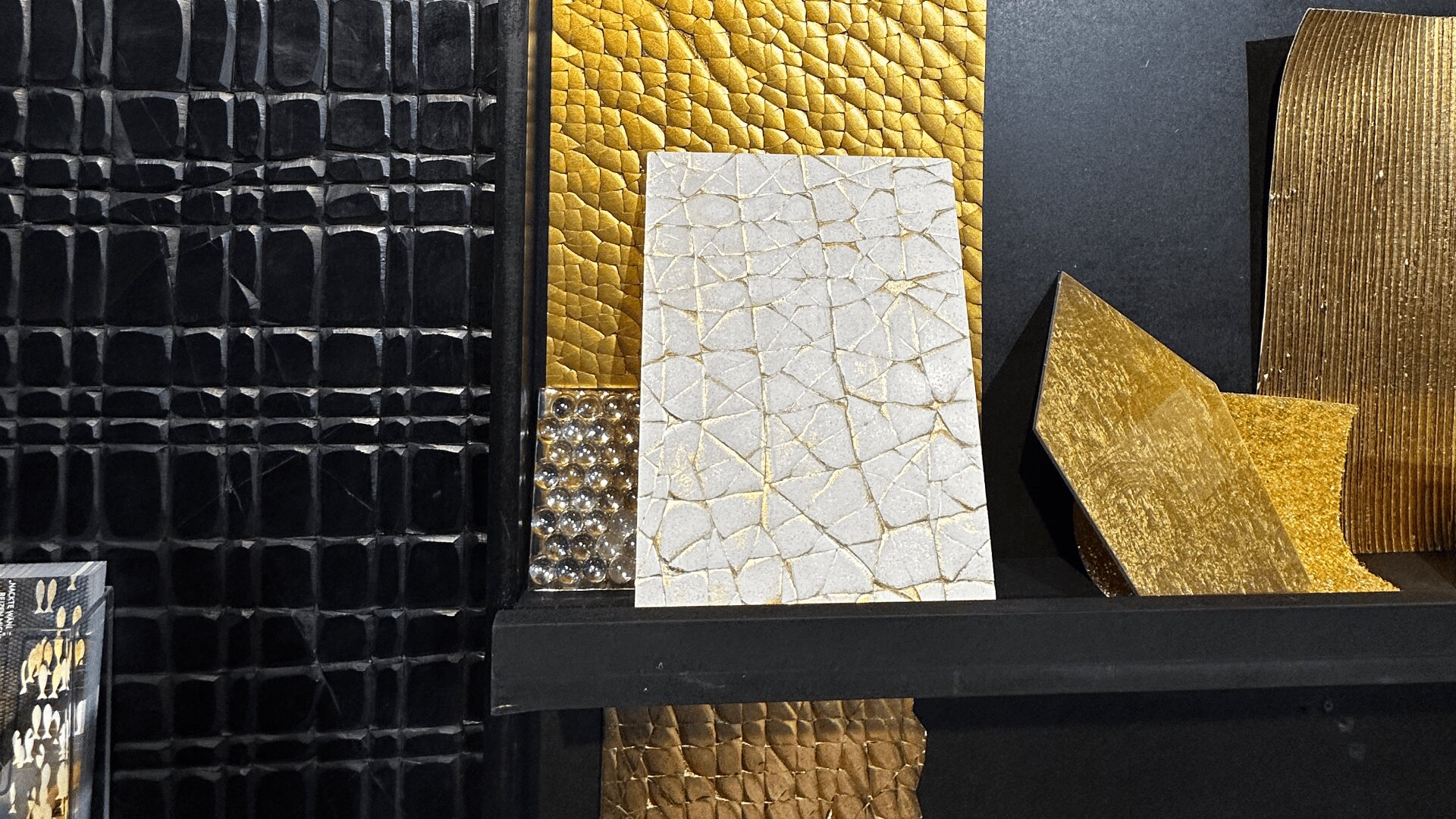 A display of textured material samples, including black, gold, and white panels with cracked, crocodile, and metallic patterns, arranged vertically and horizontally on a dark shelf.