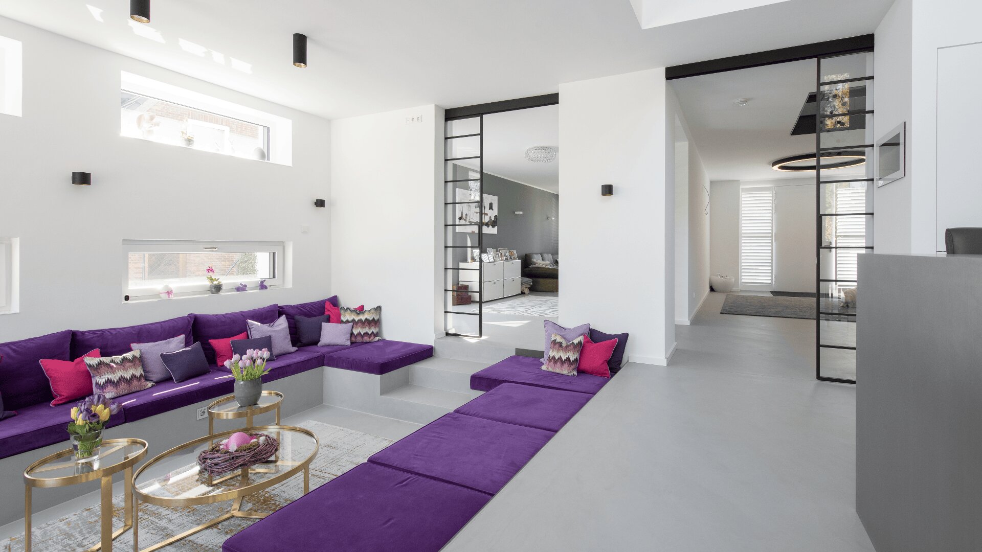 Modernes Wohnzimmer mit einem tiefliegenden lilafarbenen Sofa, bunten Kissen, goldenen runden Couchtischen und minimalistischem Dekor. Große Fenster und Glastüren lassen viel Tageslicht herein.