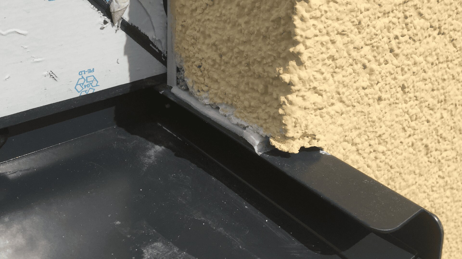Nahaufnahme einer Metallverkleidung, die an einer strukturierten, gelben Stuckwand angebracht ist. Zur Abdichtung wurde zwischen Verkleidung und Wand ein Streifen weißen Dichtmittels aufgetragen.