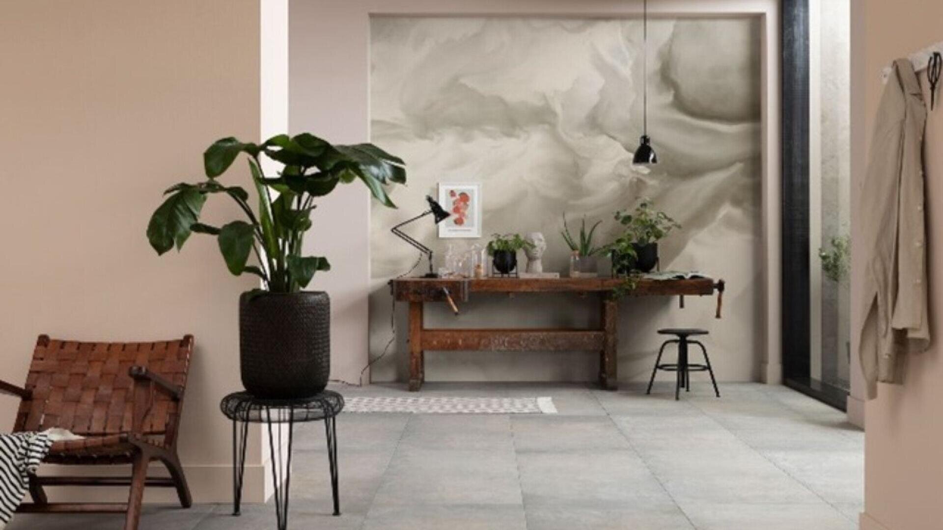 A stylish, minimalist room with a large potted plant, a woven leather chair, a rustic wooden desk with plants and art, a black stool, and a cloudy-patterned wall. Neutral tones and natural light create a calm atmosphere.