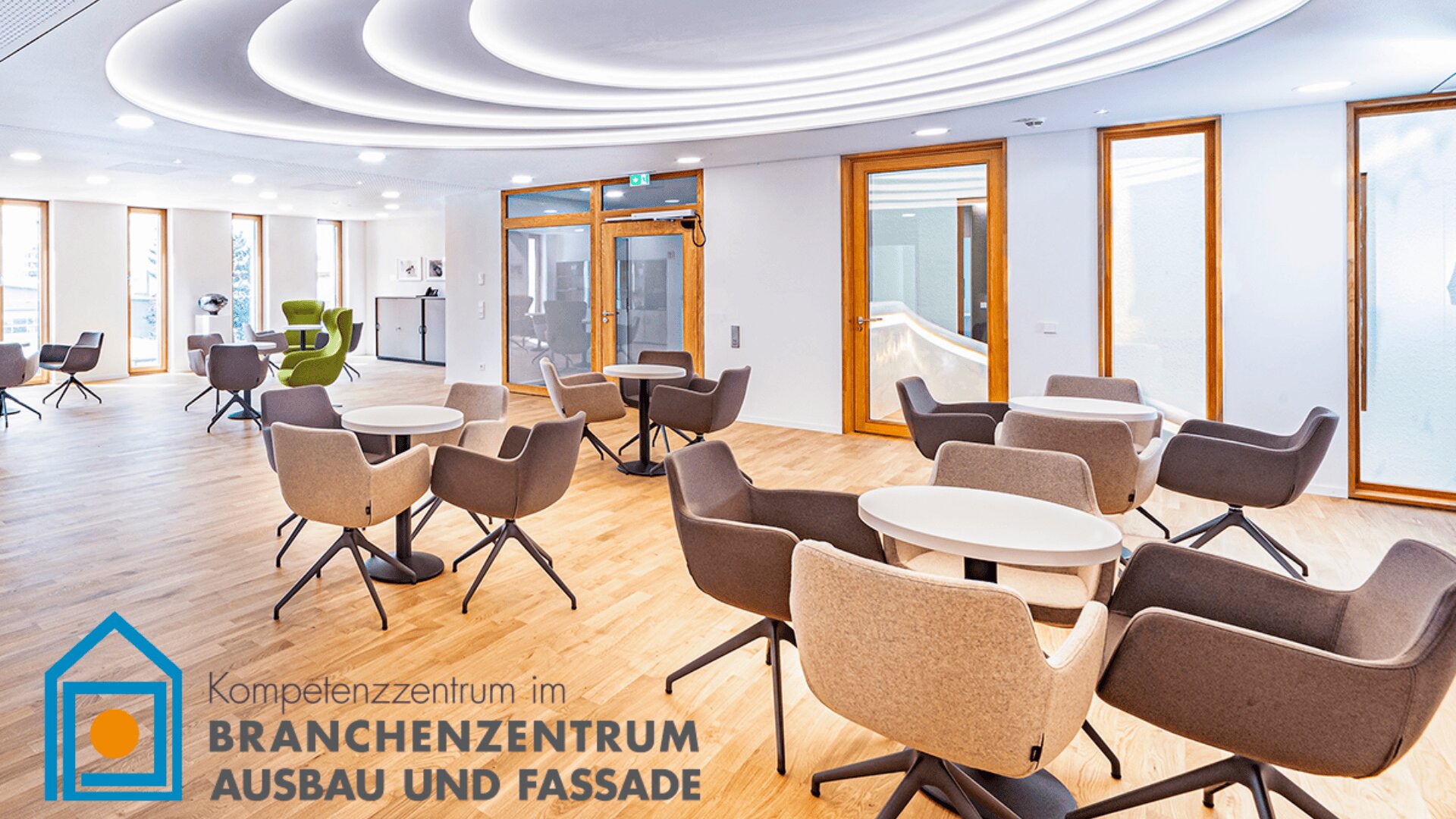 Moderner Loungebereich mit runden Tischen und grauen Stühlen auf Holzboden, großen Fenstern und Deckenleuchten. Logo und deutscher Text „Kompetenzzentrum... BRANCHENZENTRUM AUSBAU UND FASSADE“ in der unteren linken Ecke.