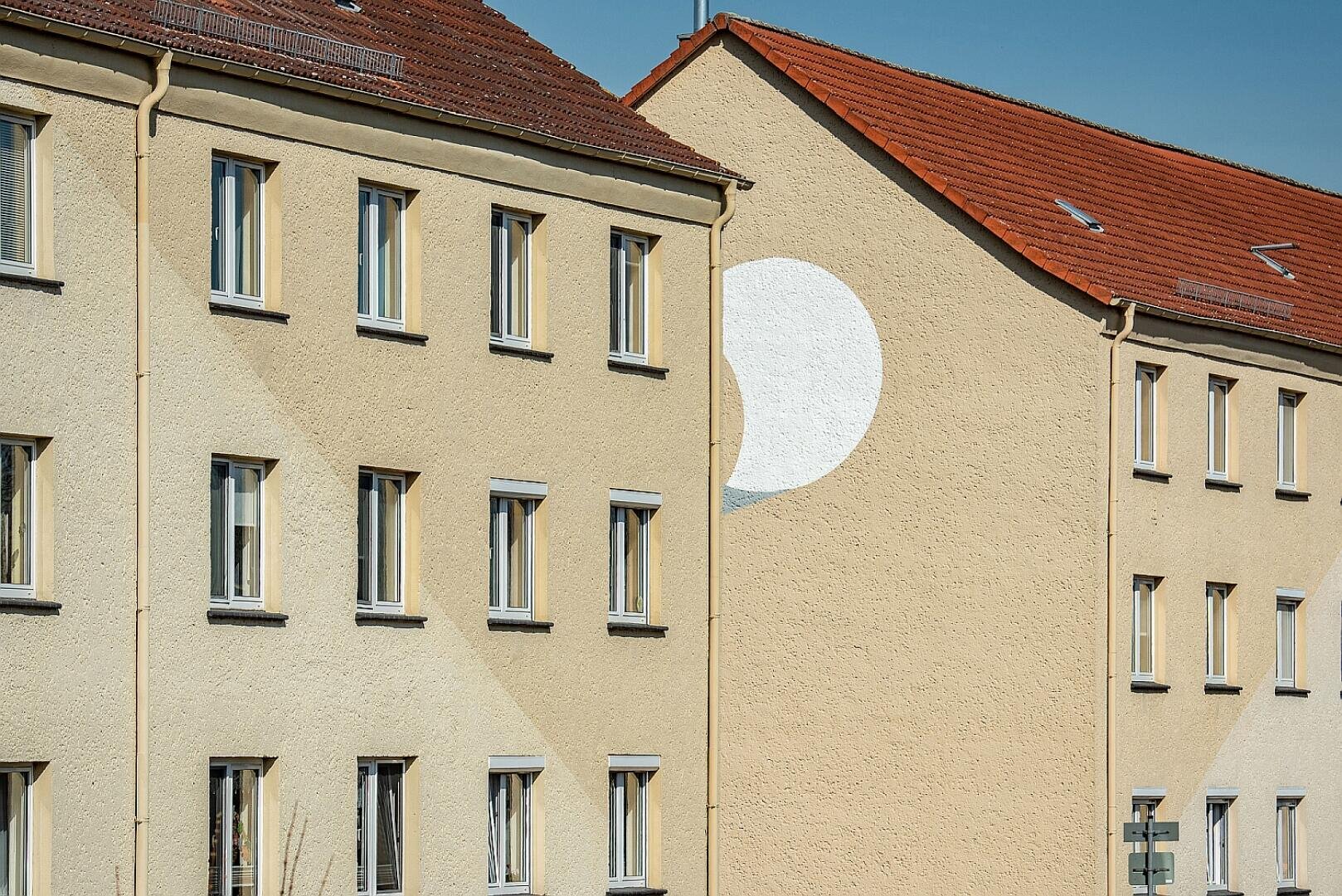 Zwei beige Wohnhäuser mit roten Ziegeldächern stehen nebeneinander. Auf die Seitenwand des rechten Gebäudes ist ein großer weißer Halbkreis gemalt, der teilweise im Schatten liegt und die Form einer auf- oder untergehenden Sonne suggeriert.