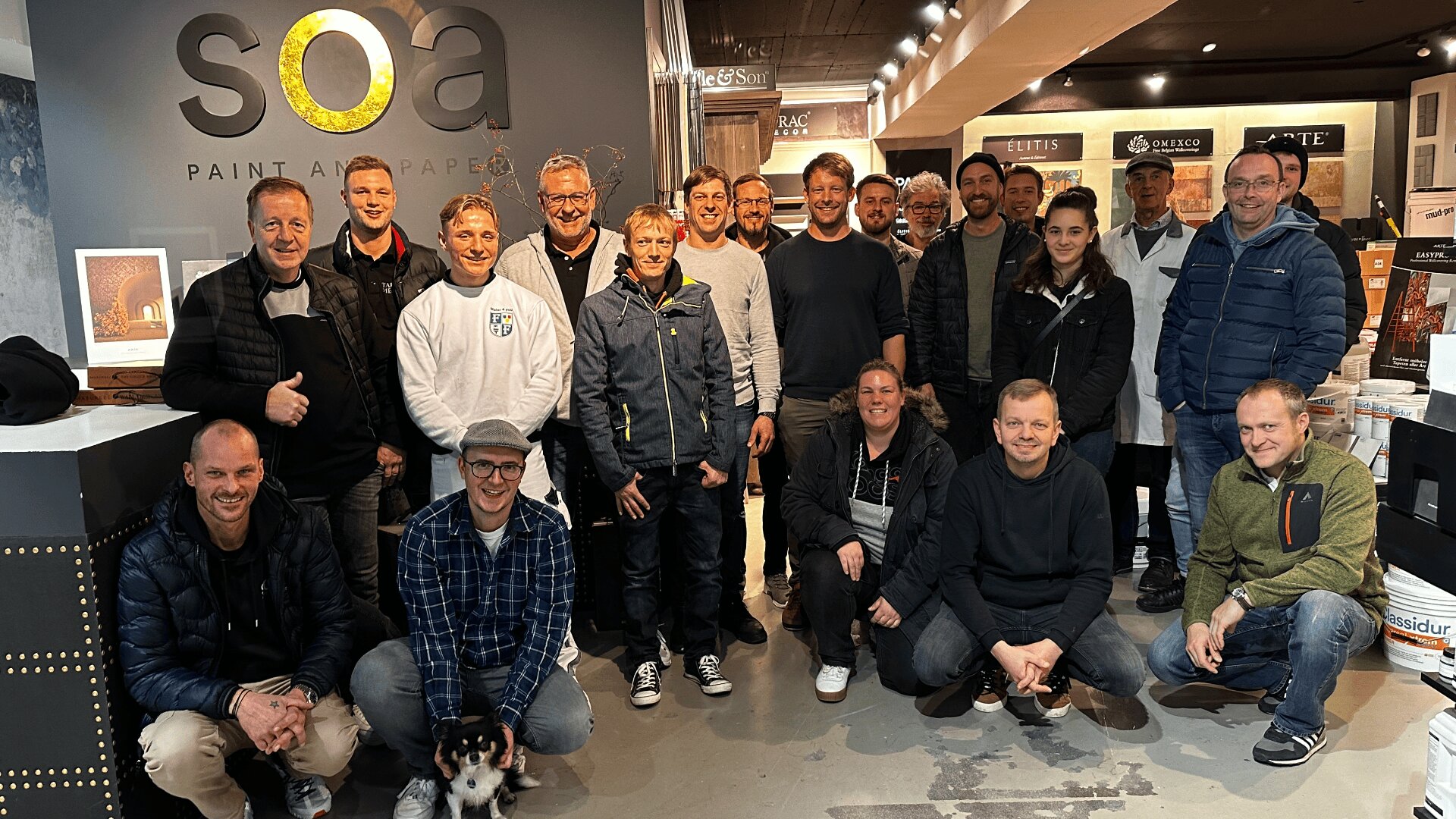Eine Gruppe von etwa 20 Personen, darunter Männer und Frauen, posiert und lächelt gemeinsam in einem modernen Ausstellungsraum mit Farb- und Tapetendisplays und dem Schild „SOA PAINT AND PAPER“ an der Wand hinter ihnen.