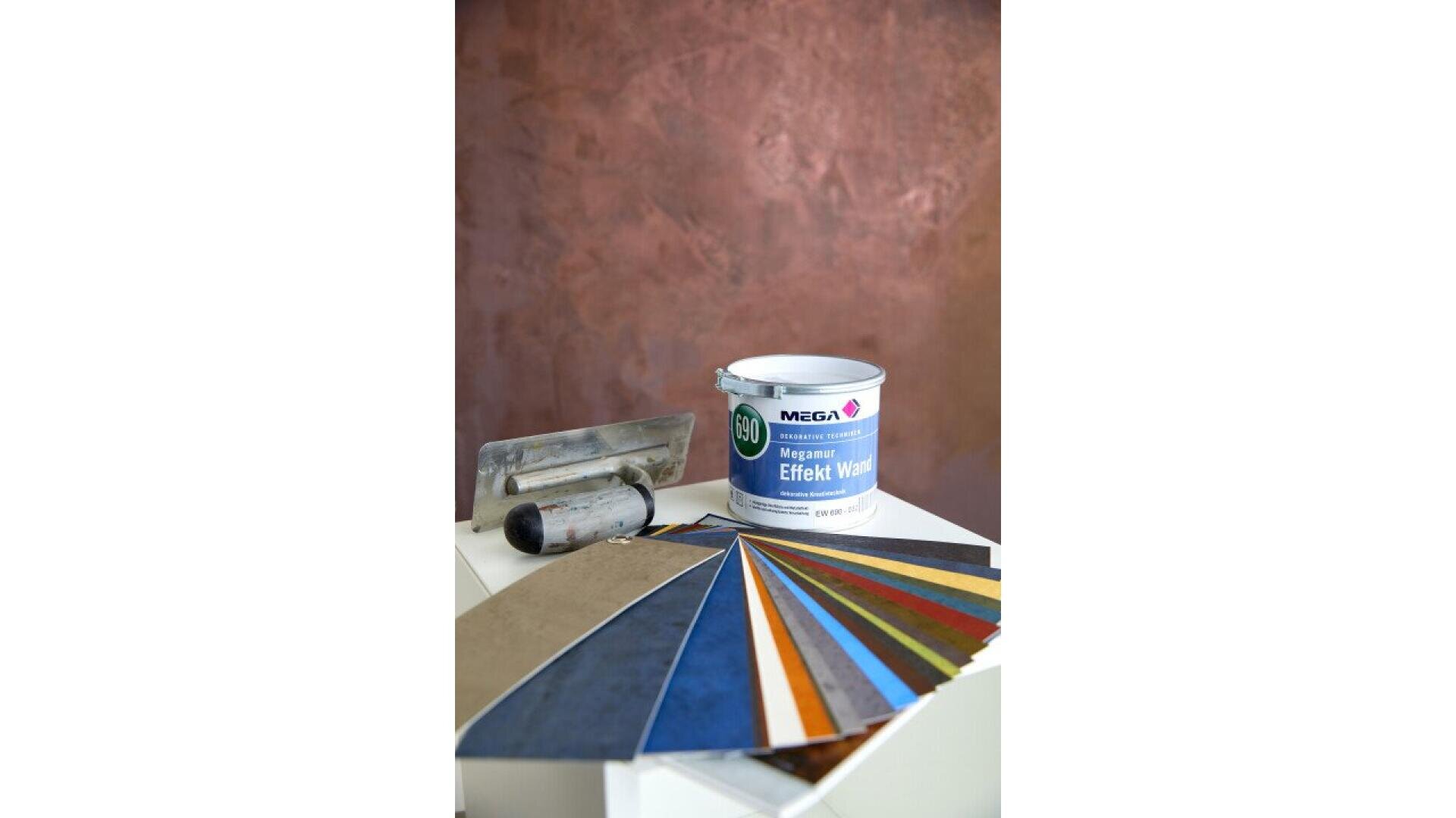A can of MEGA Effekt Wand wall paint, a metal trowel, and a fan of colorful paint samples are displayed on a white surface, with a textured brown wall in the background.