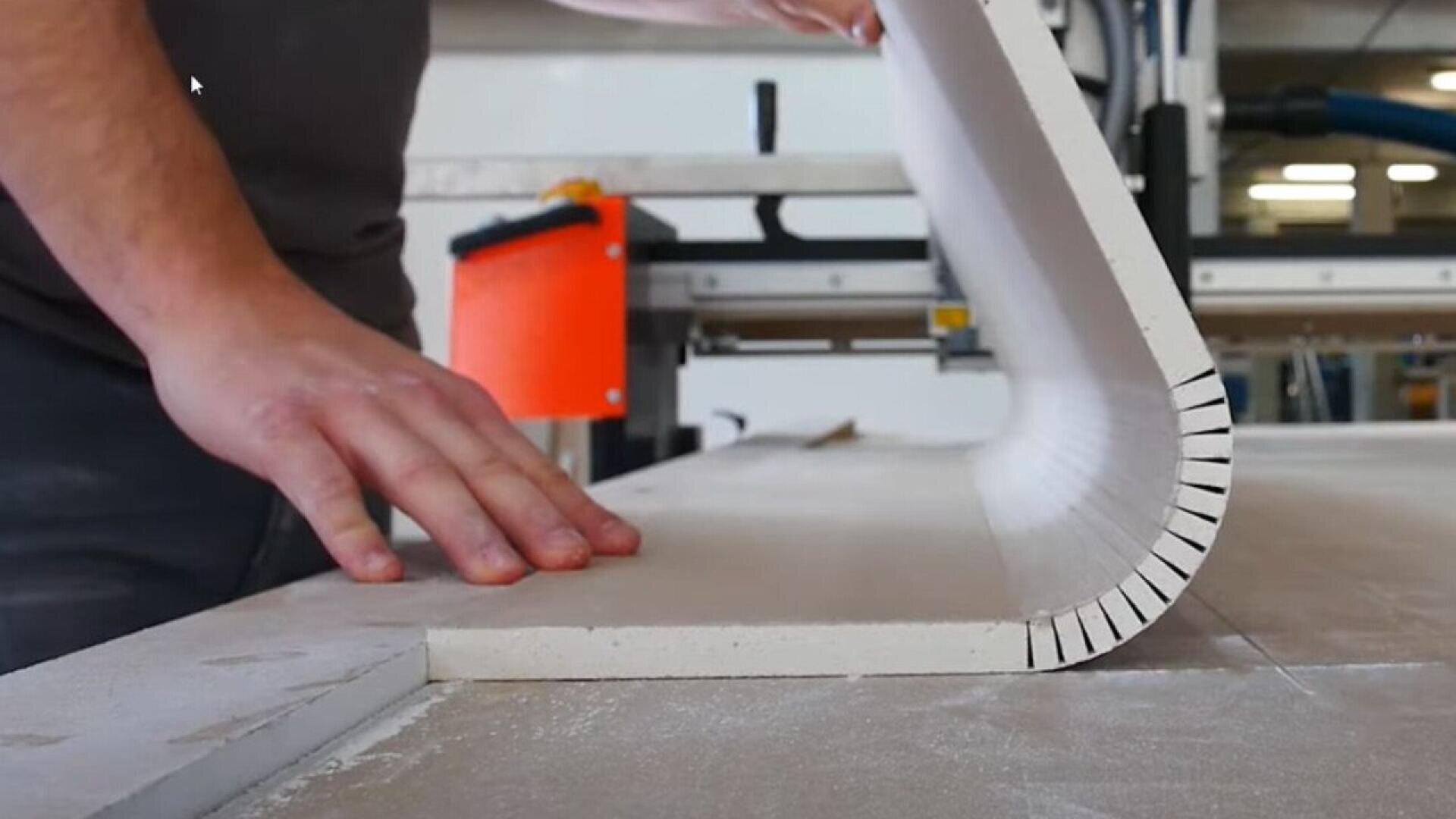 A person bends a piece of drywall with evenly spaced cuts along one side, demonstrating a technique for shaping the material on a workbench in a workshop.