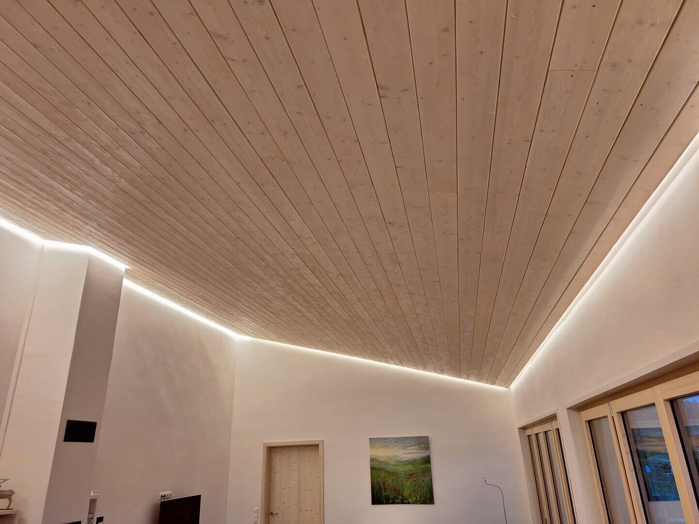 Ein moderner Raum mit einer schrägen, hellen Holzdielendecke, die durch verdeckte LED-Lichtbänder an den Rändern beleuchtet wird. Die Wände sind weiß, mit großen Fenstern und einem Landschaftsgemälde an einer Wand.