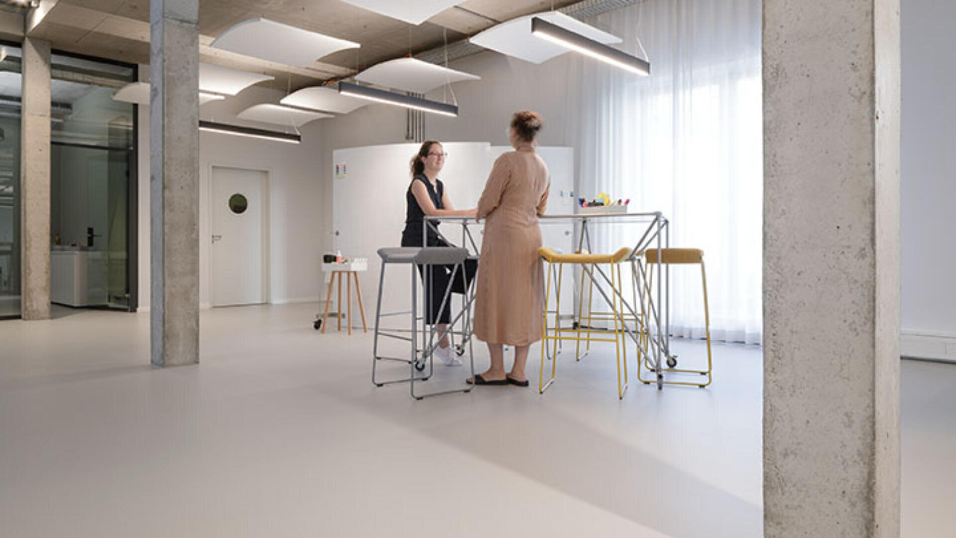 Zwei Frauen stehen und unterhalten sich an einem hohen Tisch mit gelben Hockern in einem modernen, minimalistischen Büroraum mit großen Fenstern, Betonsäulen und weißen Wänden.