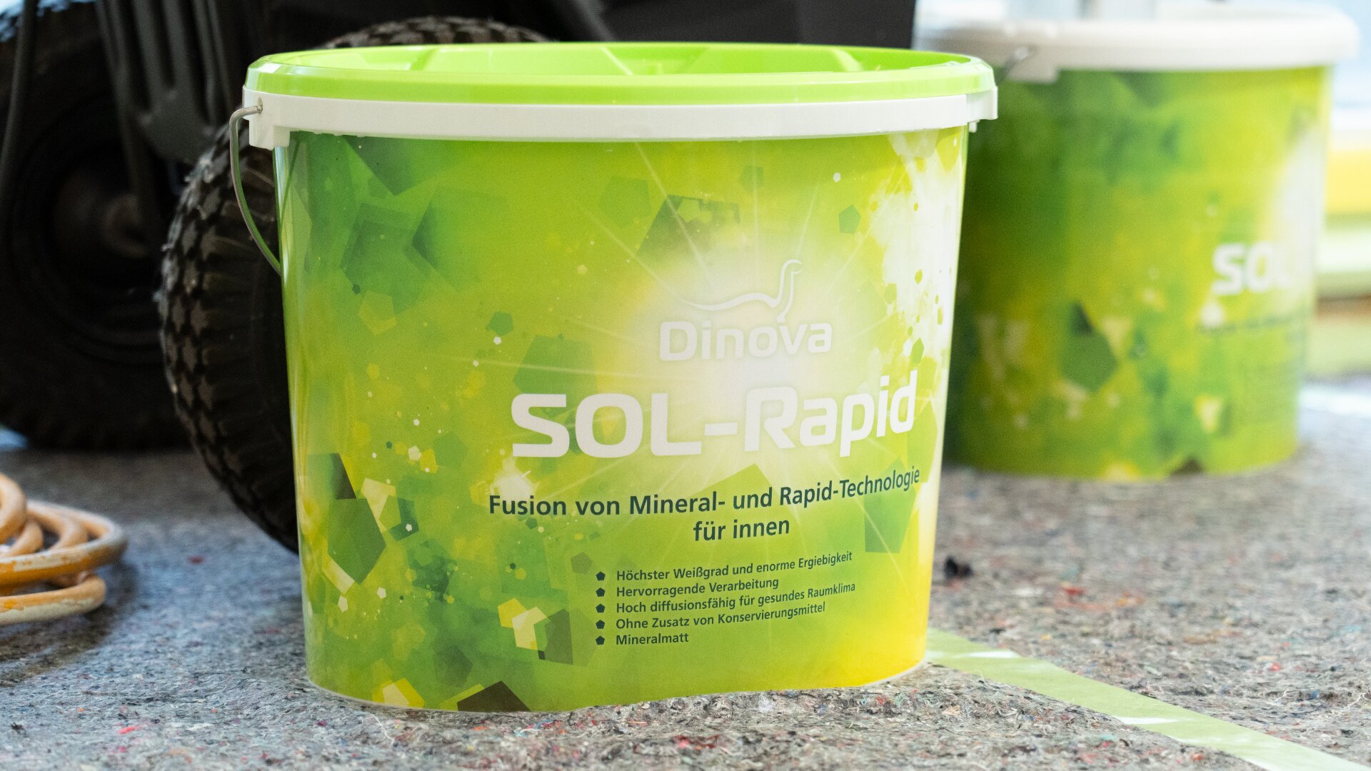 Ein grün-weißer Eimer mit der Aufschrift „Dinova SOL-Rapid“ steht auf einer gesprenkelten Fläche, im Hintergrund ein weiterer ähnlicher Eimer. Das Etikett erwähnt Mineral- und Schnelltechnologie für den Innenbereich und enthält deutschen Text.