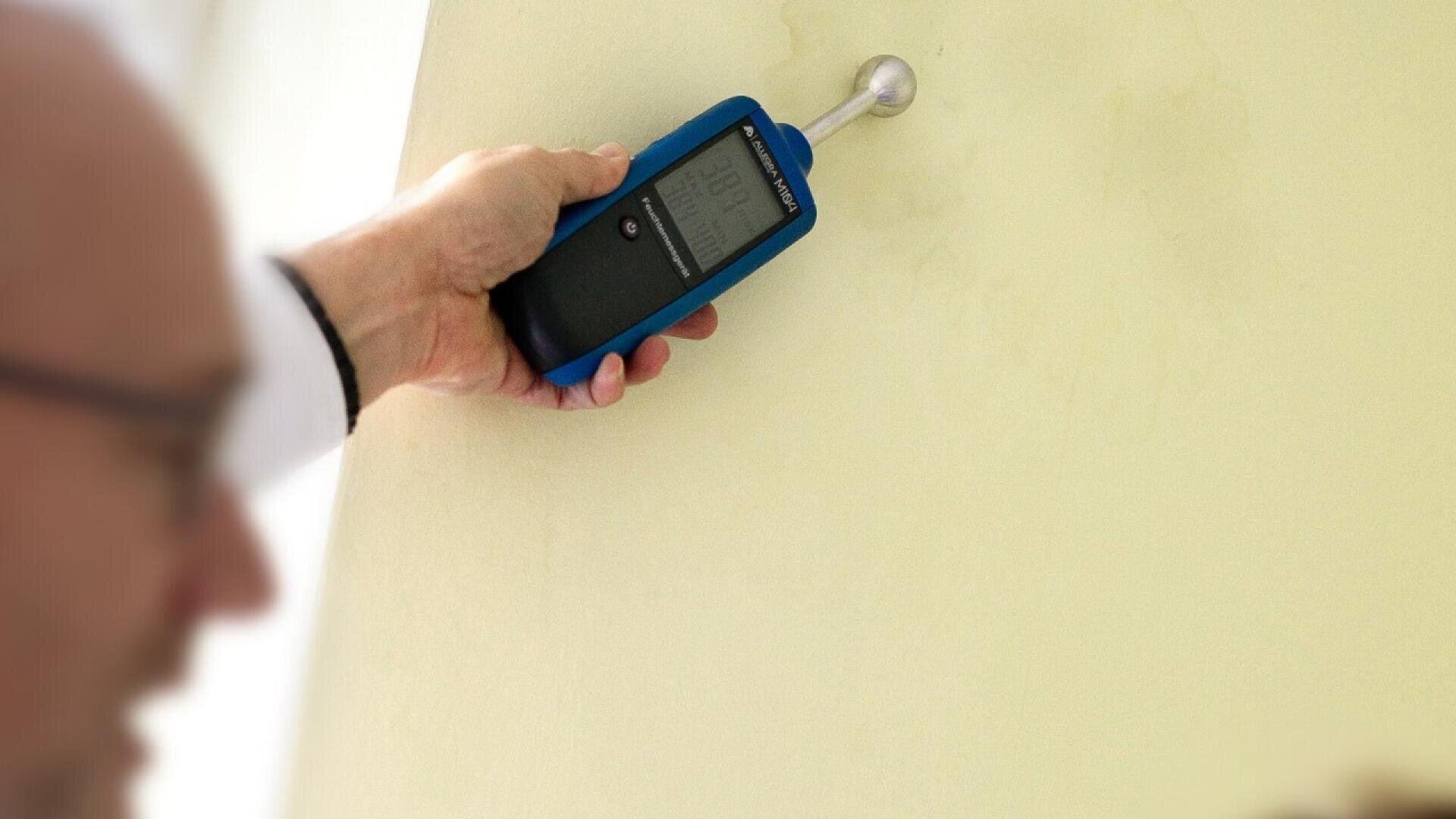 A person holds a digital moisture meter against a yellow wall, checking for dampness or moisture levels. The device displays readings on a small screen. The person’s face is partially visible and blurred in the foreground.