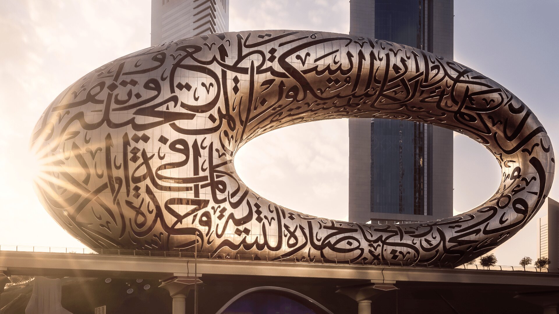 A futuristic oval building covered in Arabic calligraphy stands against the skyline at sunset, with sunlight shining through its central opening.