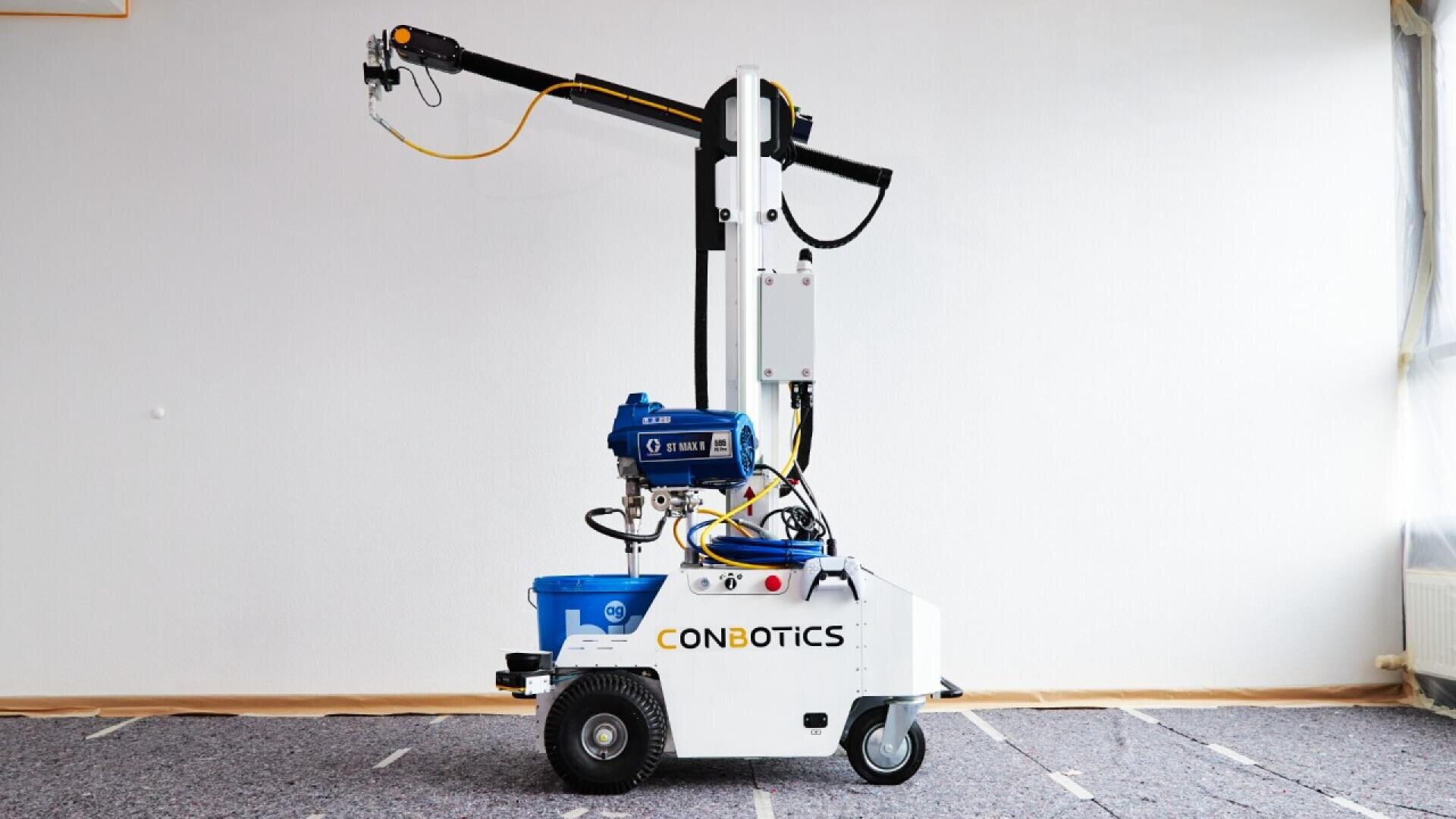 A white robotic machine labeled CONBOTICS with wheels, a blue component, and an extendable arm, stands on a carpeted floor in a plain, partially-lit room.