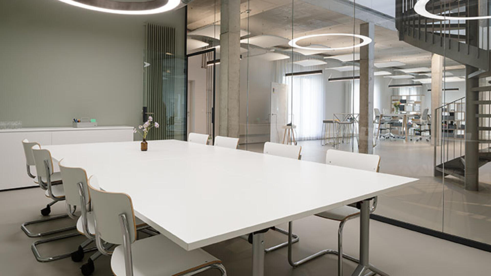 Moderner Konferenzraum mit großem weißen Tisch, acht weißen Stühlen, minimalistischem Dekor, Glaswänden und einer Wendeltreppe im Hintergrund. Kreisförmige Deckenleuchten sorgen für Beleuchtung.
