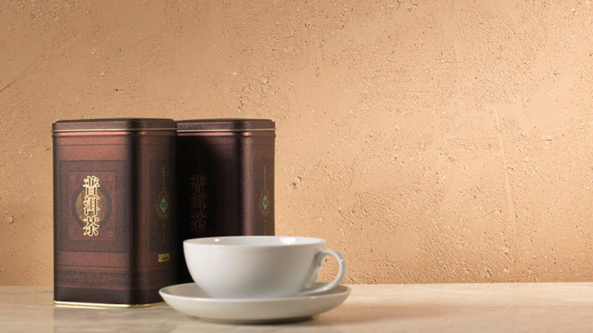 A white teacup and saucer sit on a light-colored surface next to two brown tea tins with gold writing, set against a textured beige wall.