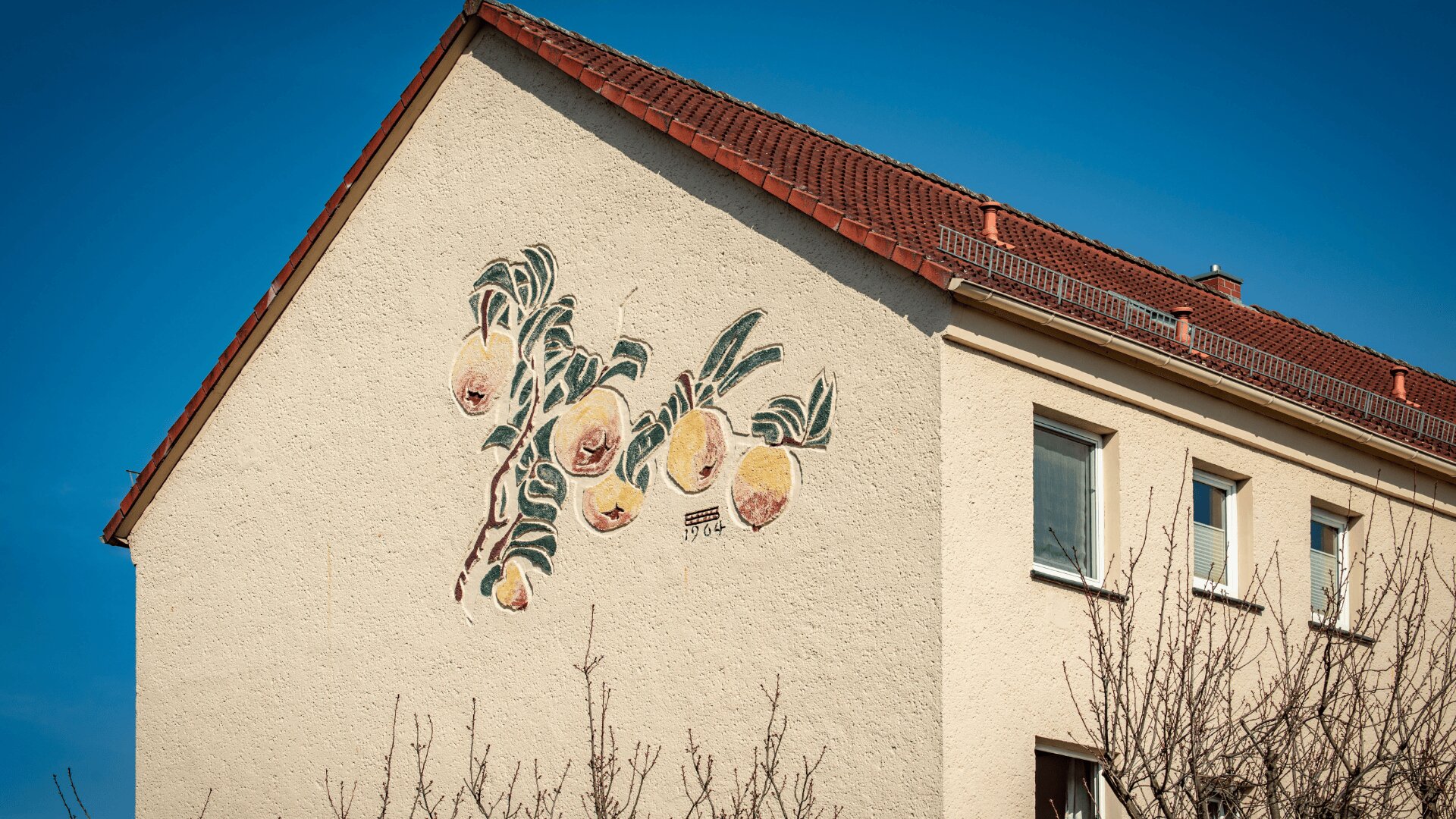 Auf die Seite eines beigen Gebäudes mit einem roten Ziegeldach unter einem klaren blauen Himmel ist ein Wandgemälde eines fruchttragenden Zweigs, wahrscheinlich mit Pfirsichen, gemalt.