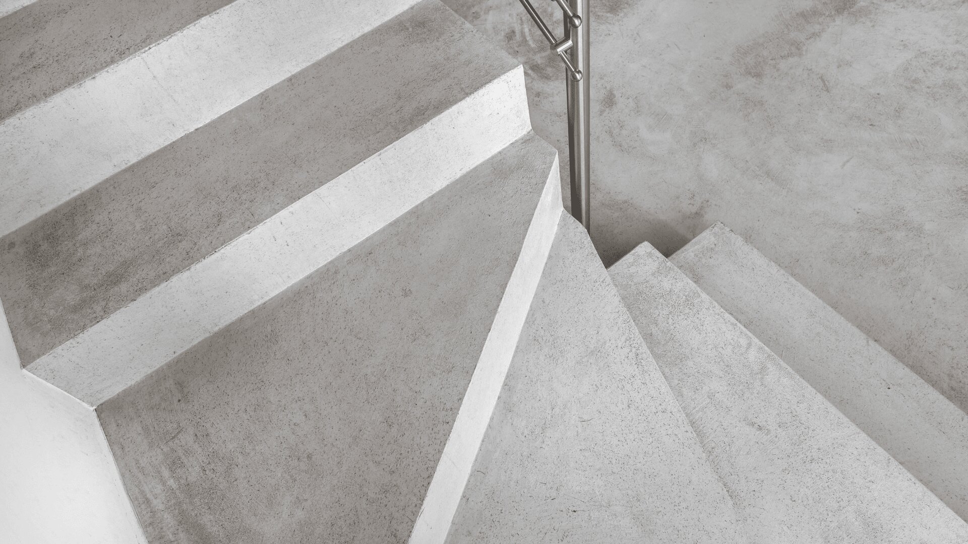 A top-down view of a minimalist concrete staircase with a smooth, gray finish and a simple metal handrail on the right side. The stairs and landing have a modern, industrial look.